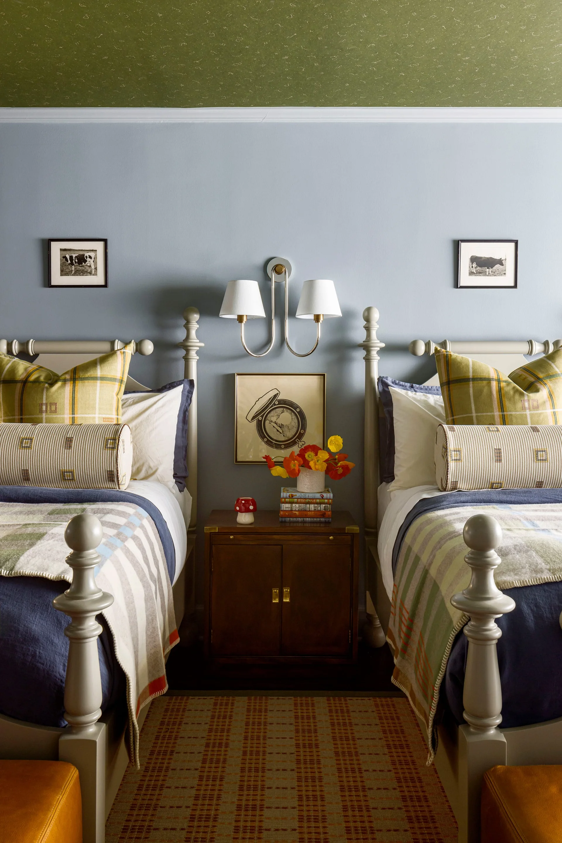 Two symmetrical twin beds with plaid pillows flank a wooden nightstand against blue walls under a green textured ceiling.