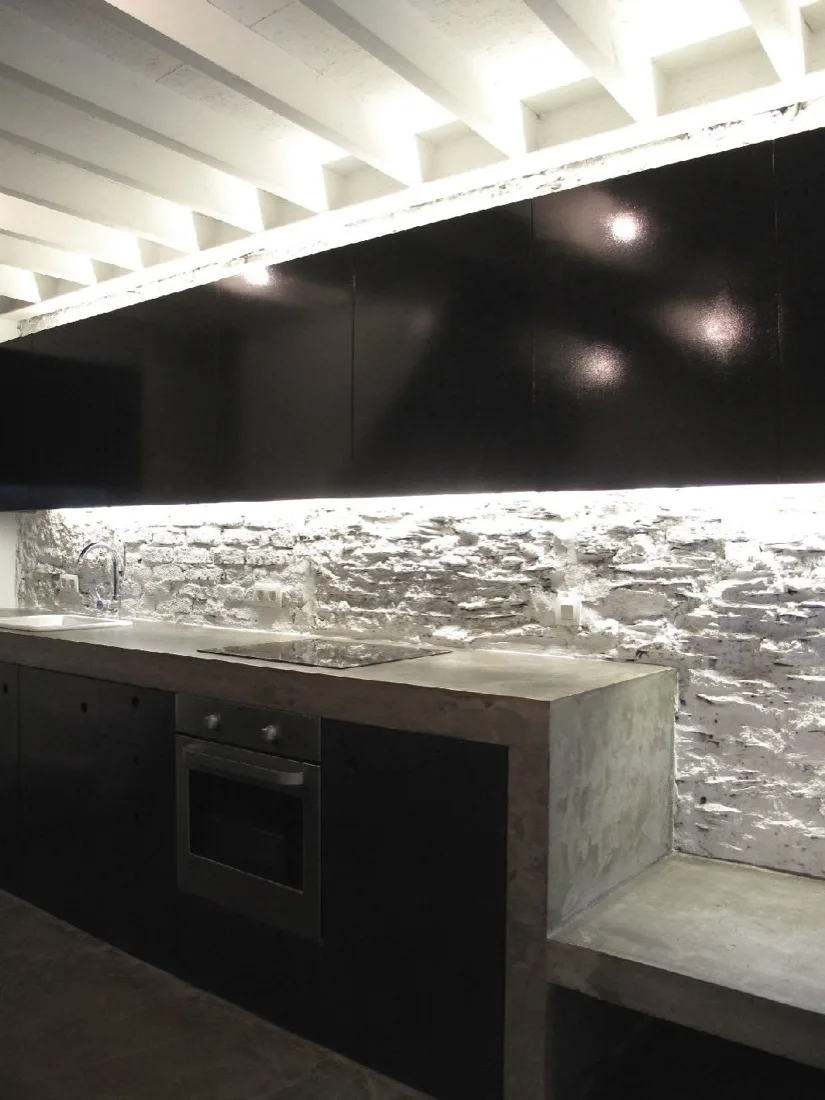 A minimalist kitchen features matte black cabinetry, a concrete countertop with a built-in oven, and a white-painted rough stone backsplash under beams.