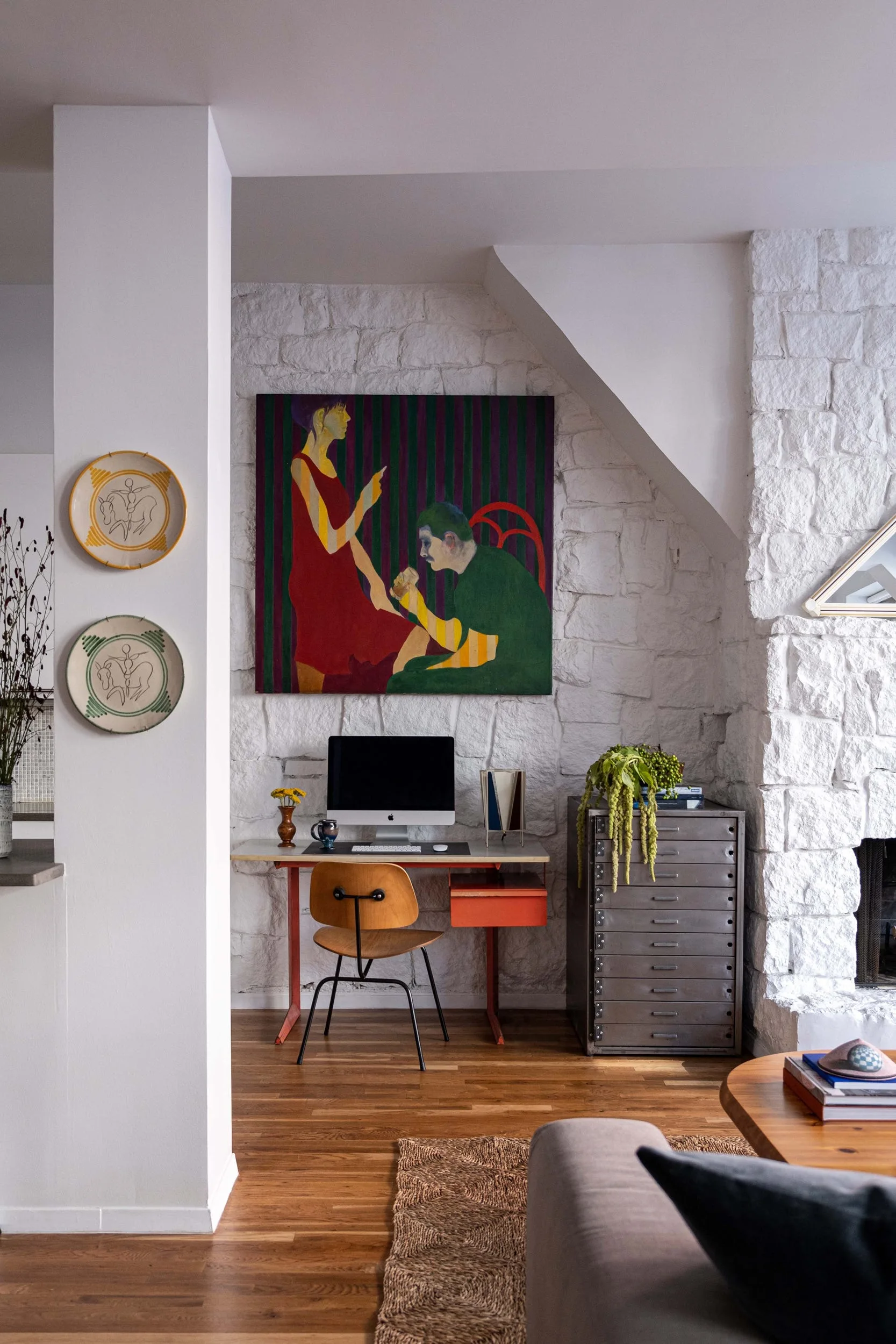 A compact home office nook features a wood desk, metal storage cabinet, and large colorful painting set against a white textured stone wall.