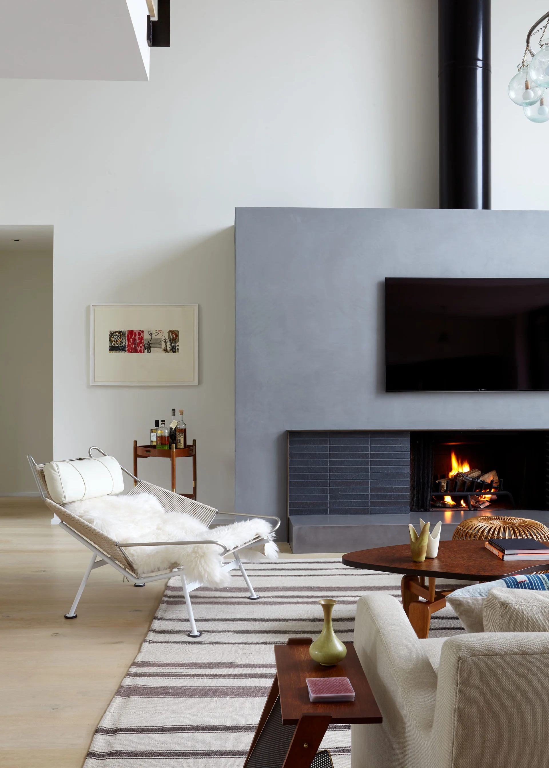 A modern living room featuring a gray plastered fireplace with a mounted TV, a metal lounge chair with a sheepskin throw, and a striped area rug.