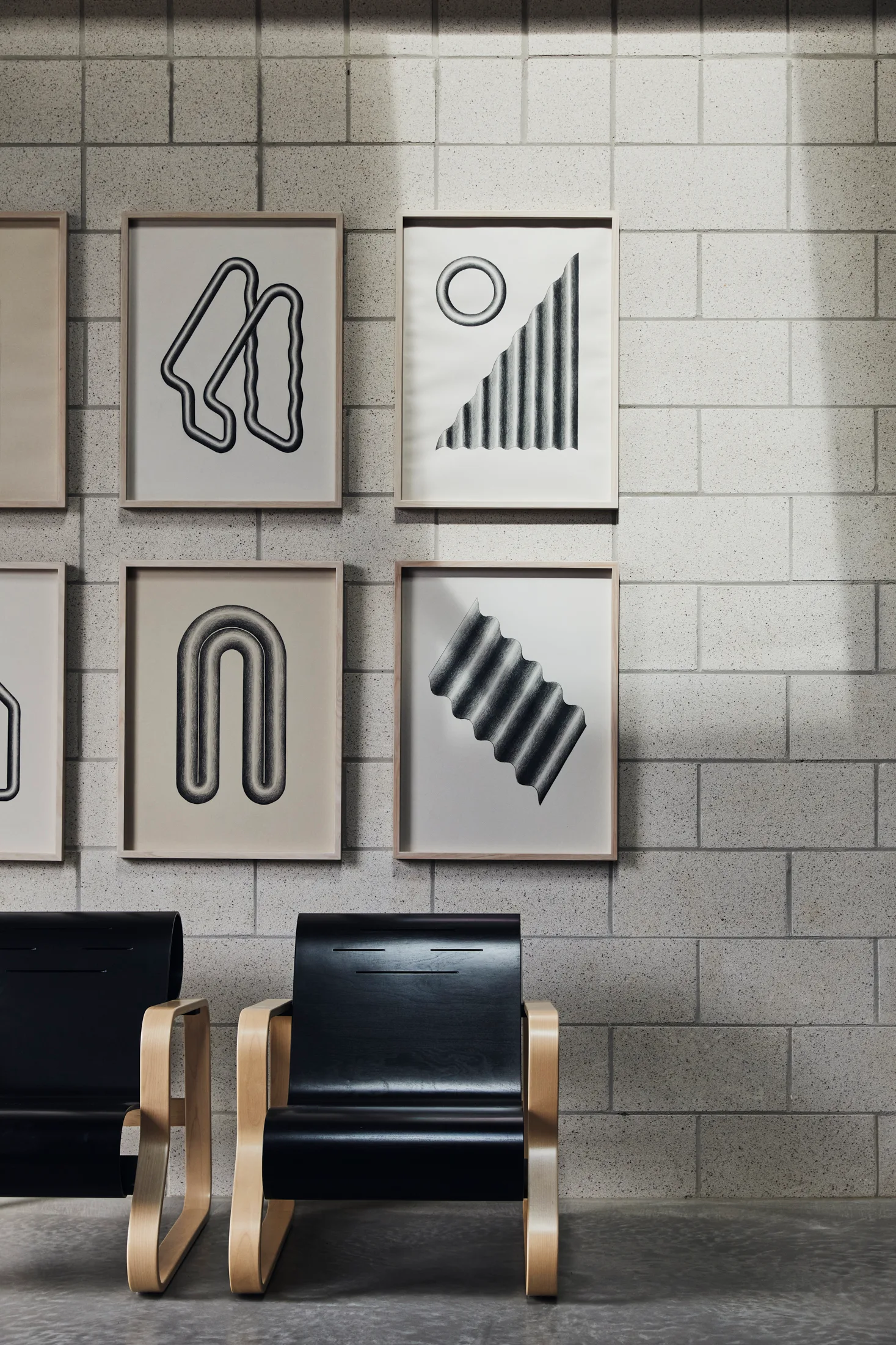 Two black bentwood armchairs sit below a grid of framed abstract art prints on a light gray concrete block wall.