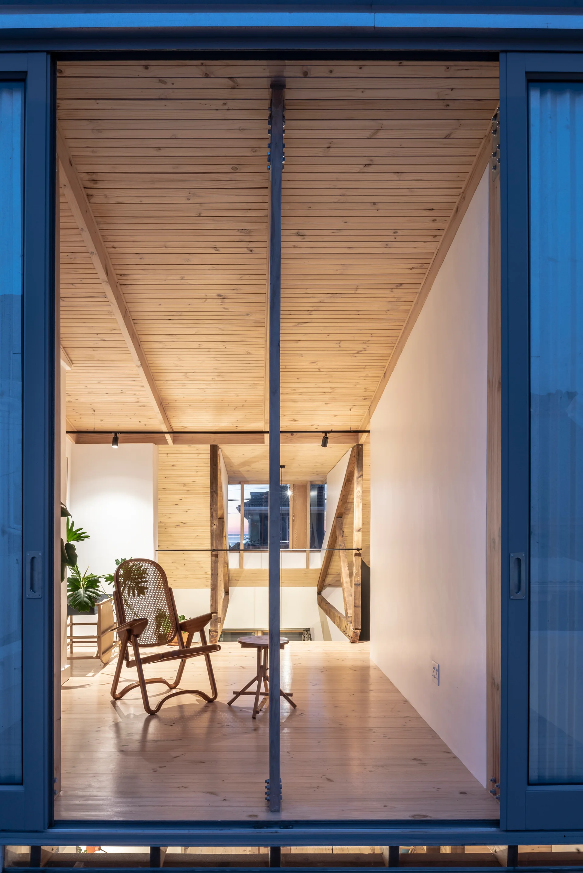 A modern loft interior viewed through sliding doors features a vaulted wood-paneled ceiling, light hardwood floors, and a cane lounge chair.