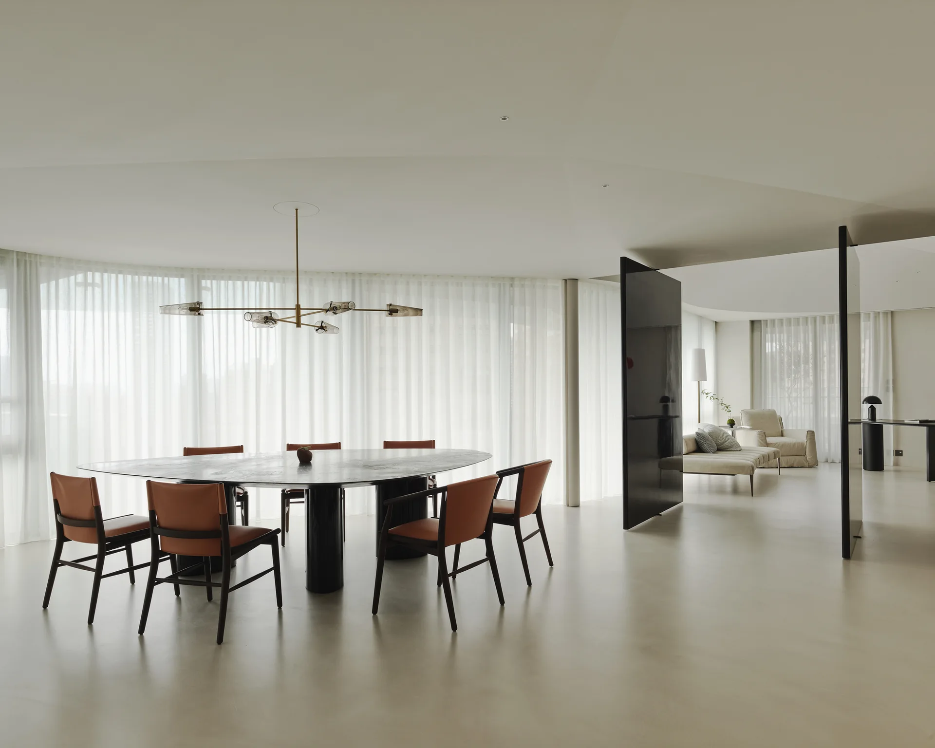 A minimalist dining room features a dark elliptical table, warm orange leather chairs, a brass branching chandelier, and sheer white curtains.