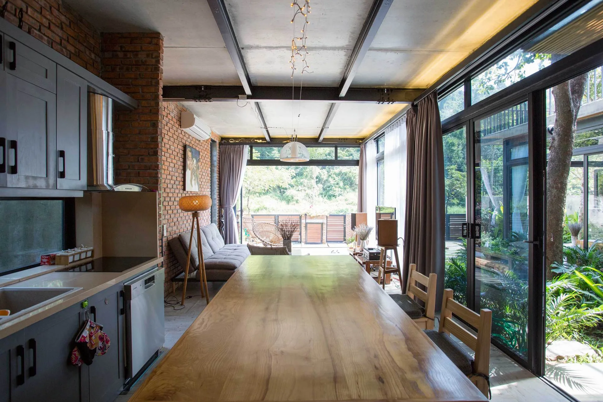 An open kitchen and dining space with a long wooden table, an exposed brick wall, dark ceiling beams, and glass doors opening to a lush garden.