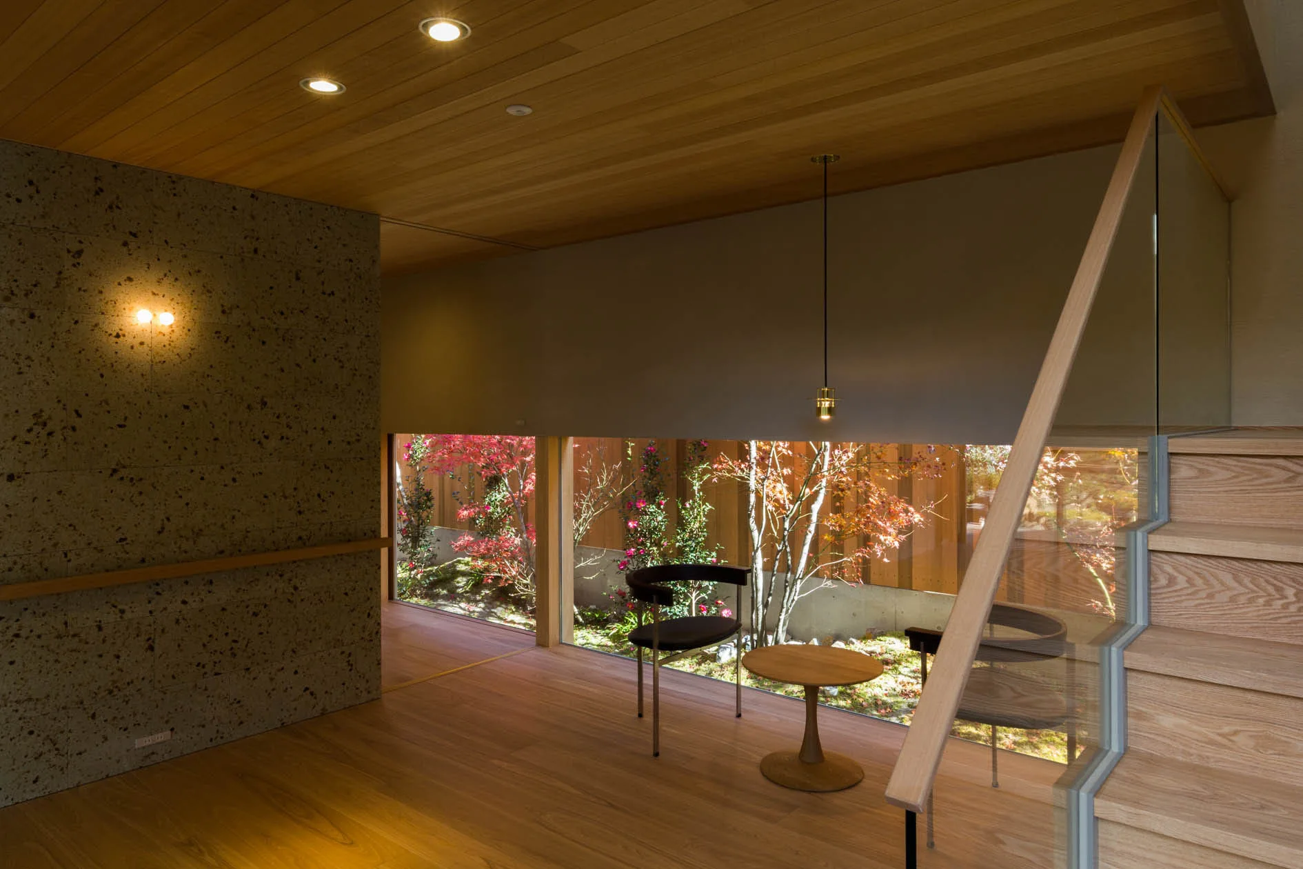A modern interior with a wooden staircase, wood slat ceiling, and a low horizontal window framing a garden with red and green plants.