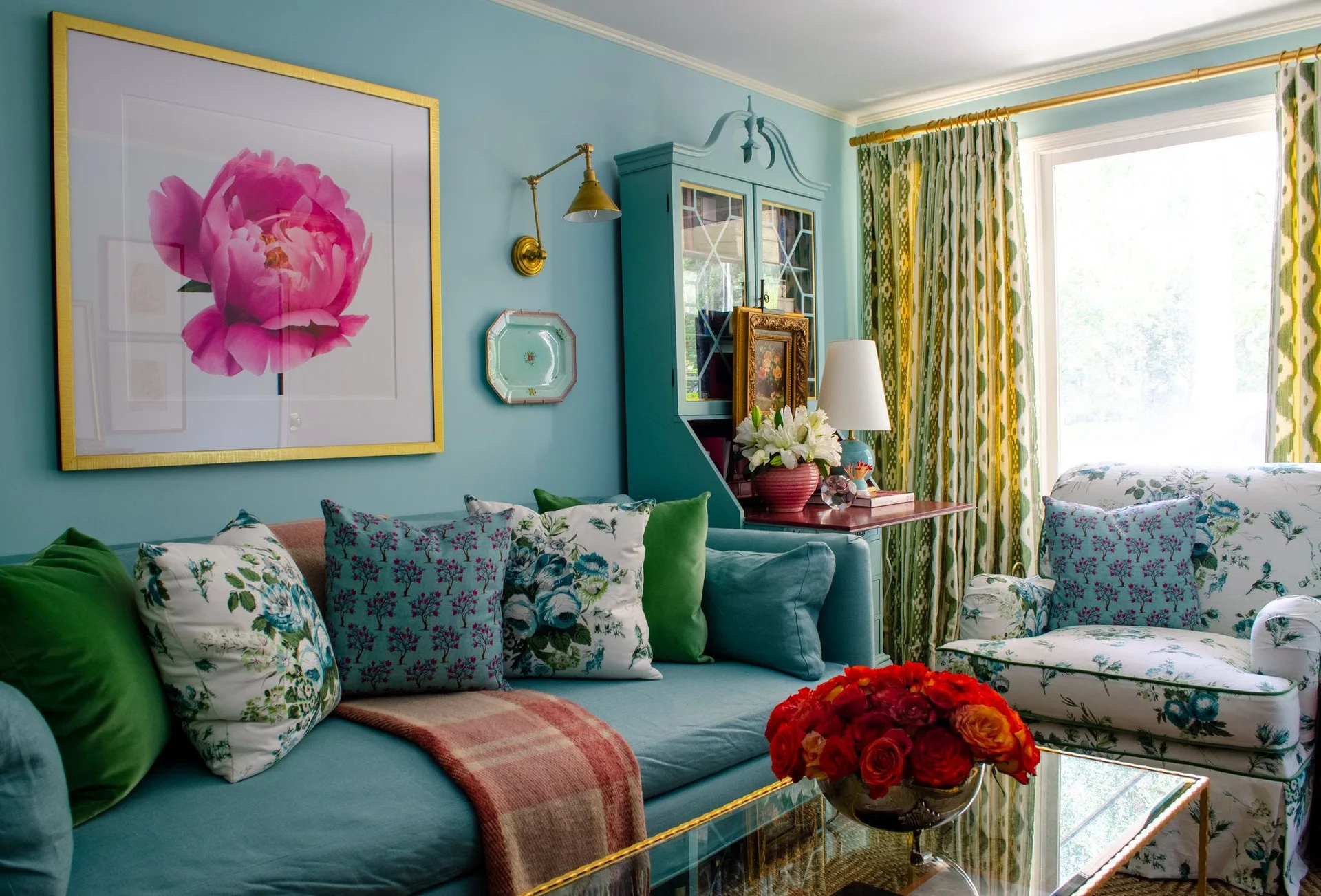 A light blue living room featuring a matching blue sofa, a painted wood secretary cabinet, patterned curtains, and a floral armchair.