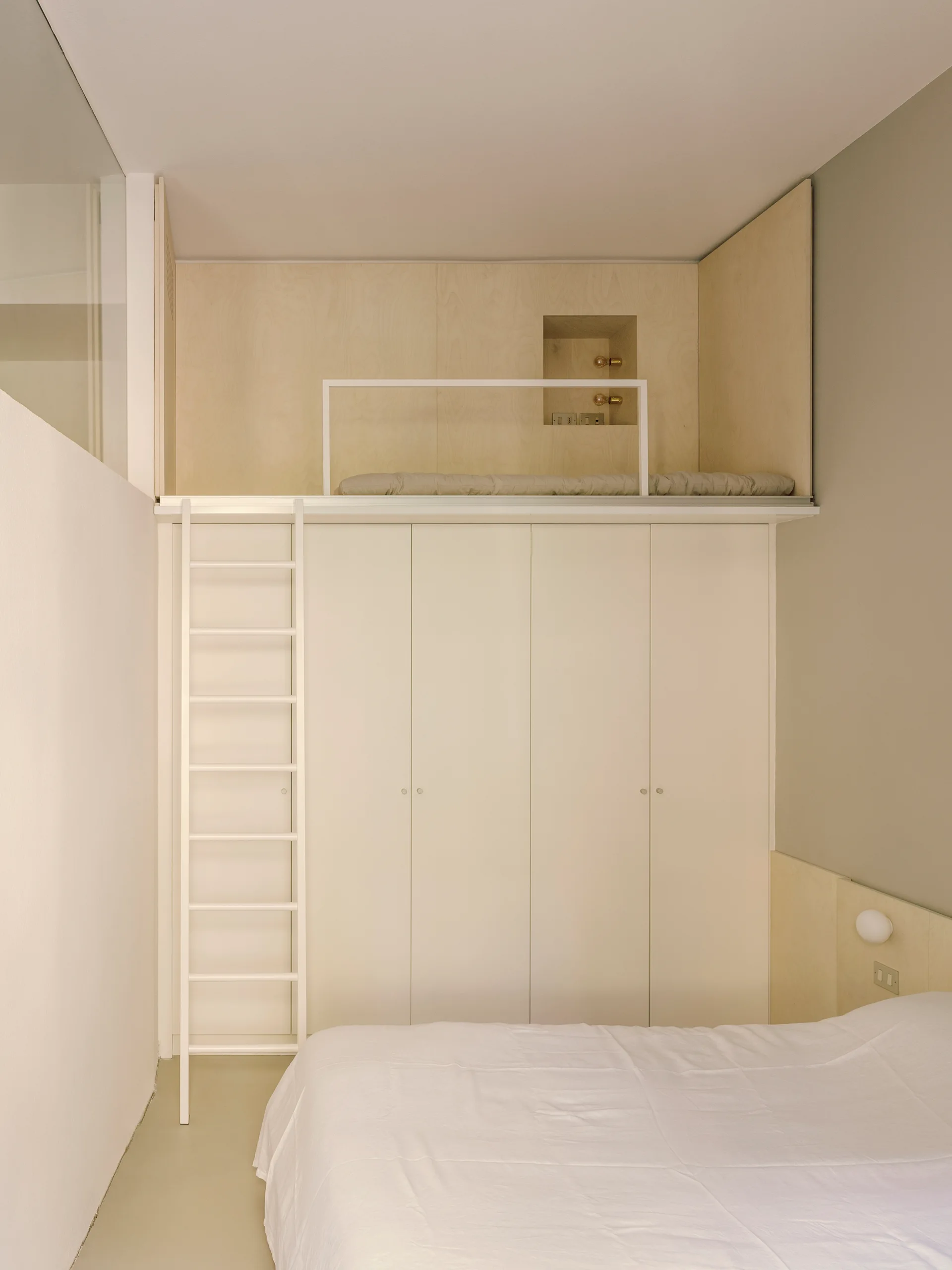 A small bedroom features a bed in the foreground and a tall white built-in wardrobe supporting an elevated plywood loft bed with a white ladder.