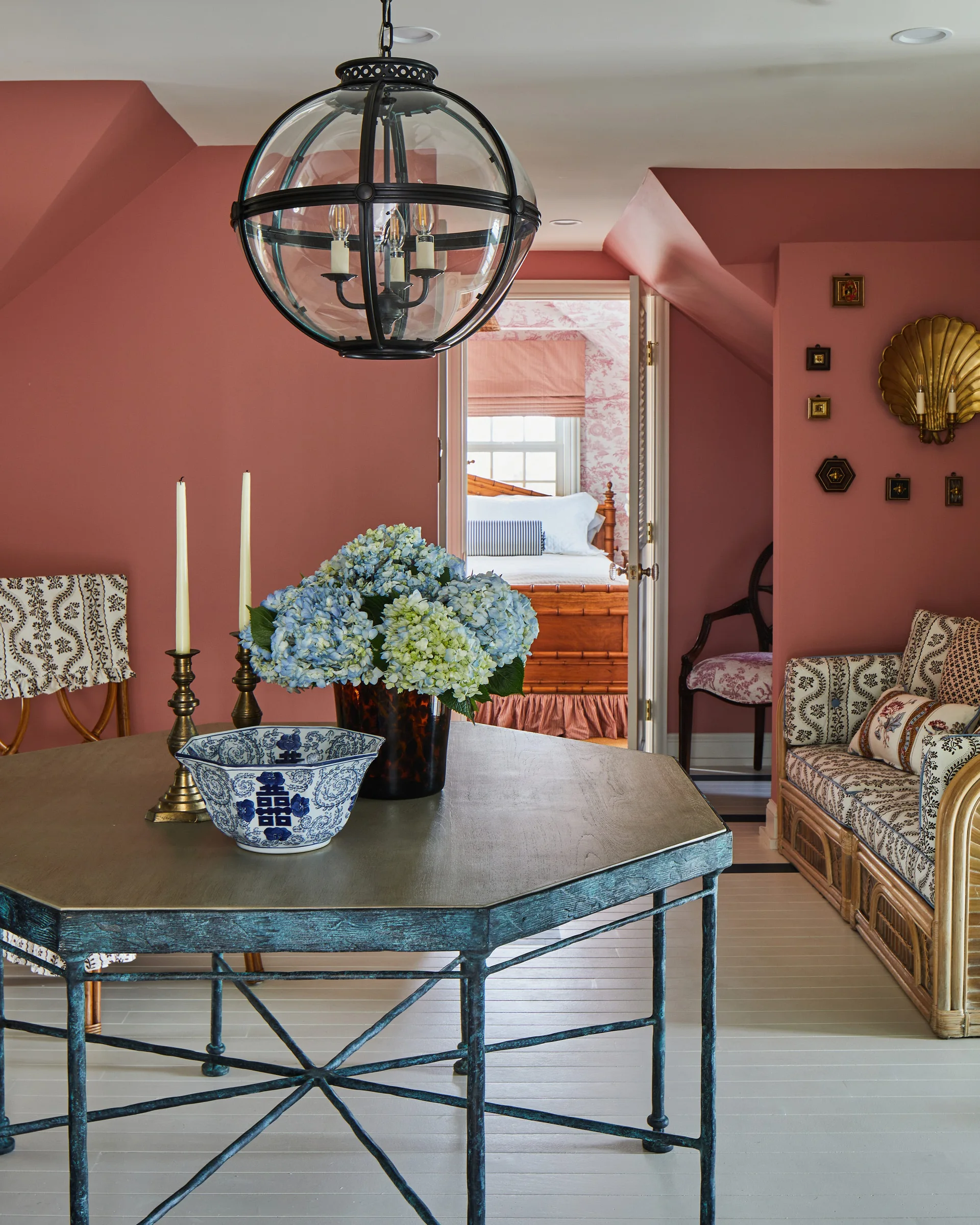 Dusty rose sitting room with a hexagonal wood table, a rattan sofa with patterned cushions, and a doorway opening to a bedroom.