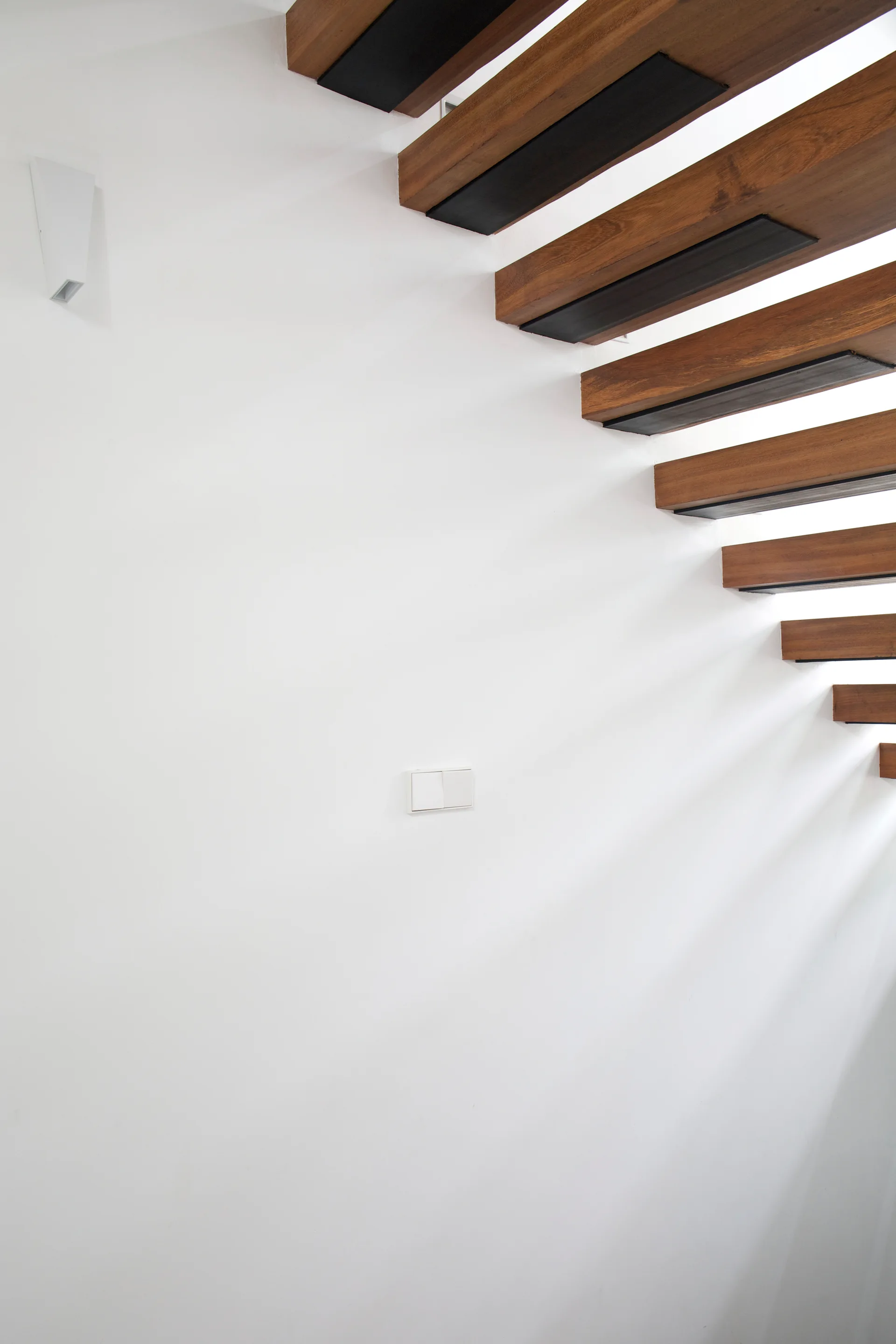 Floating wooden stair treads with dark undersides ascend against a plain white wall casting diagonal shadows.
