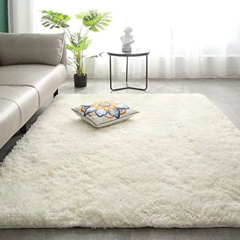 A fluffy white shag rug lies on a grey floor next to a light grey sofa, a small black side table, and a potted plant in front of a window.