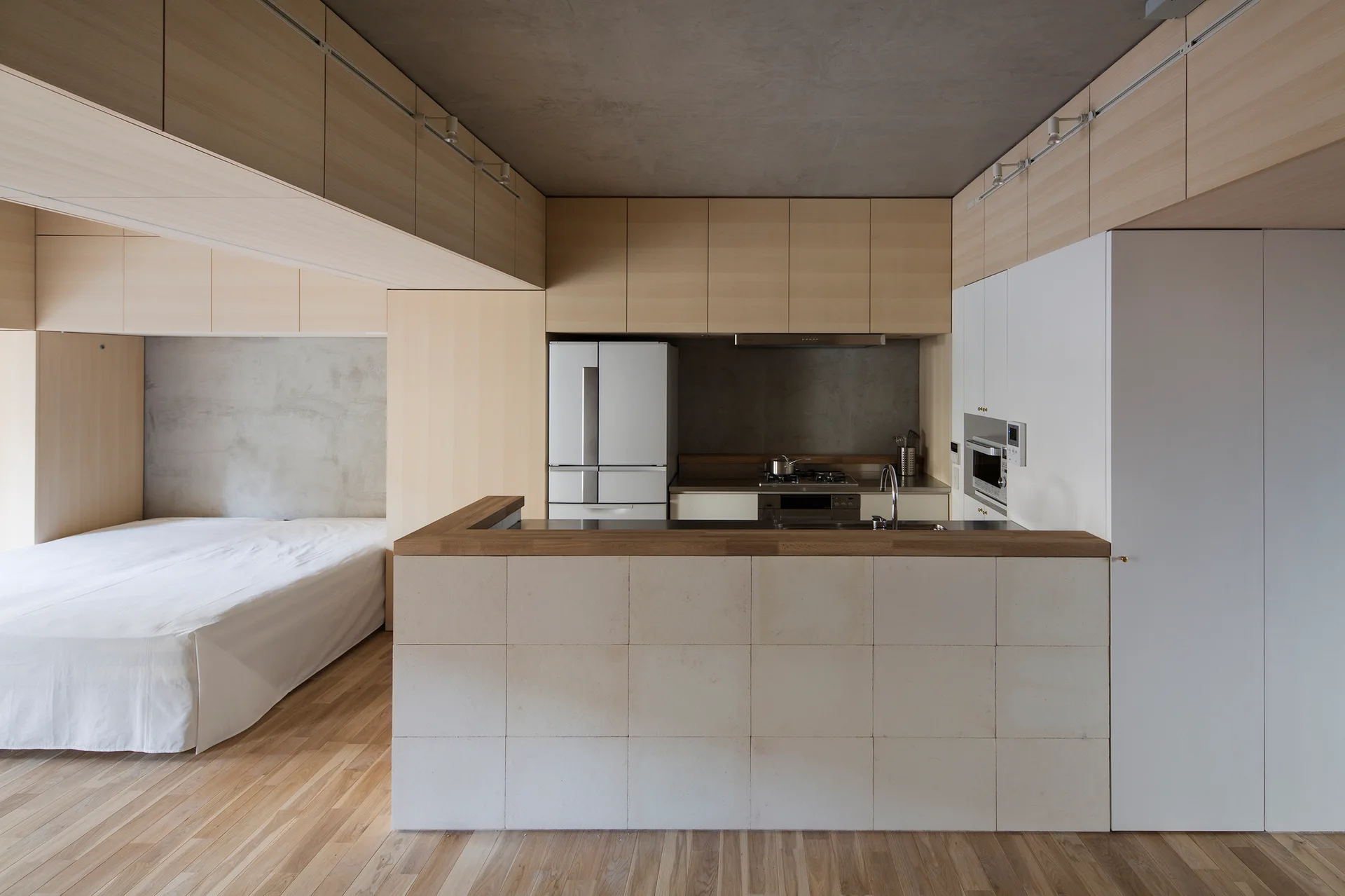 A minimalist studio apartment features a kitchen with light wood cabinets and a stone-tiled island next to a bed area under a concrete ceiling.