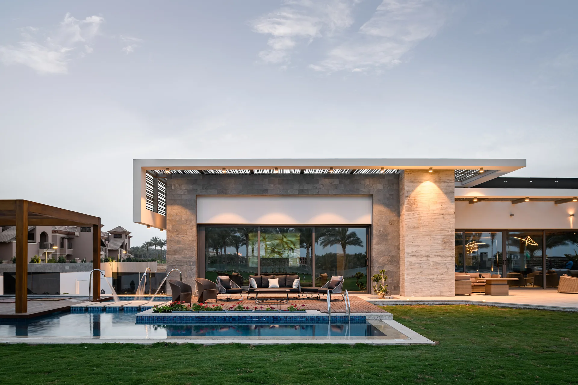 A contemporary backyard patio features a swimming pool with fountains, a wooden deck with dark outdoor furniture, and a stone-clad exterior.