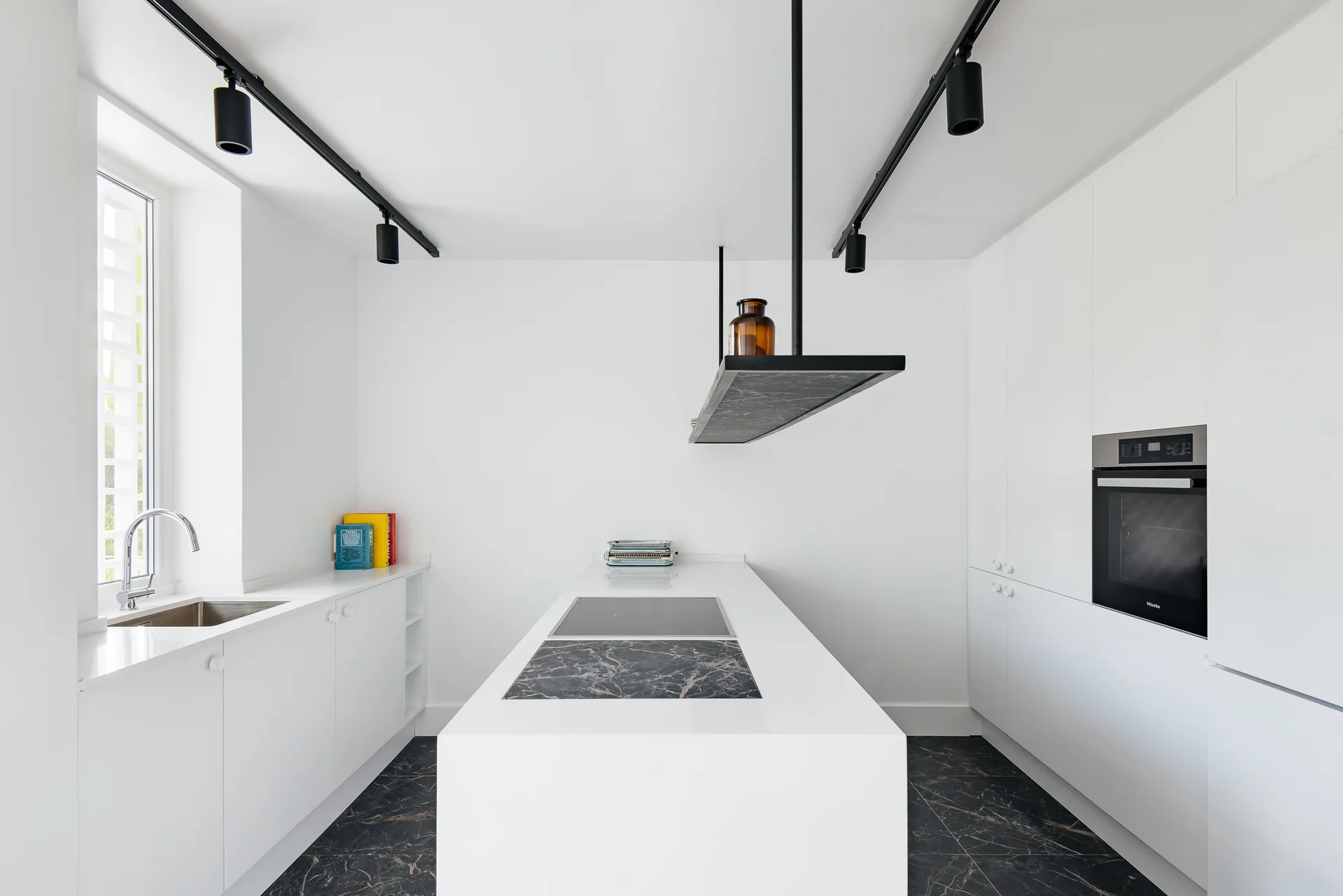 A minimalist white kitchen features a central island with a marbled stone inlay, dark patterned stone flooring, and black ceiling track lights.