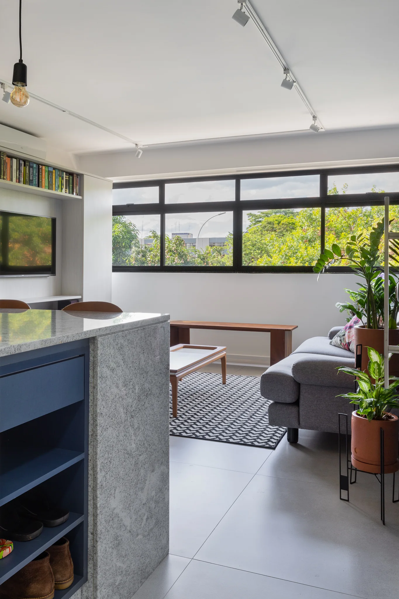 A modern open-plan living room features a grey sofa, geometric area rug, wide window, and a stone-topped kitchen island with blue shelving in the foreground.