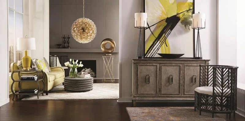 A transitional living room featuring a yellow sofa, a round grey ottoman, and a rustic wooden console table under a large abstract painting.