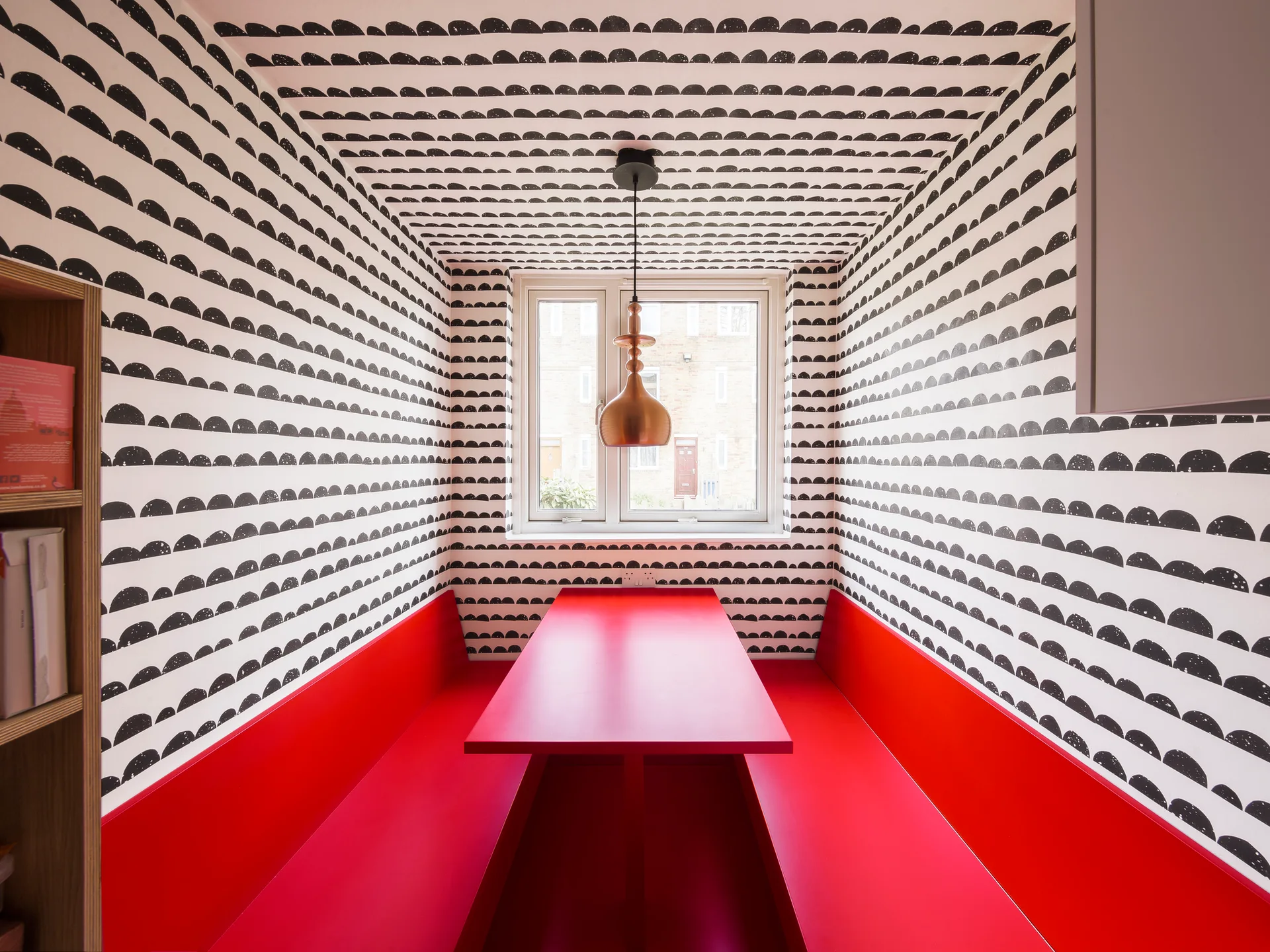 A compact dining nook features a bright red table and bench seating surrounded by black and white patterned wallpaper on the walls and ceiling.