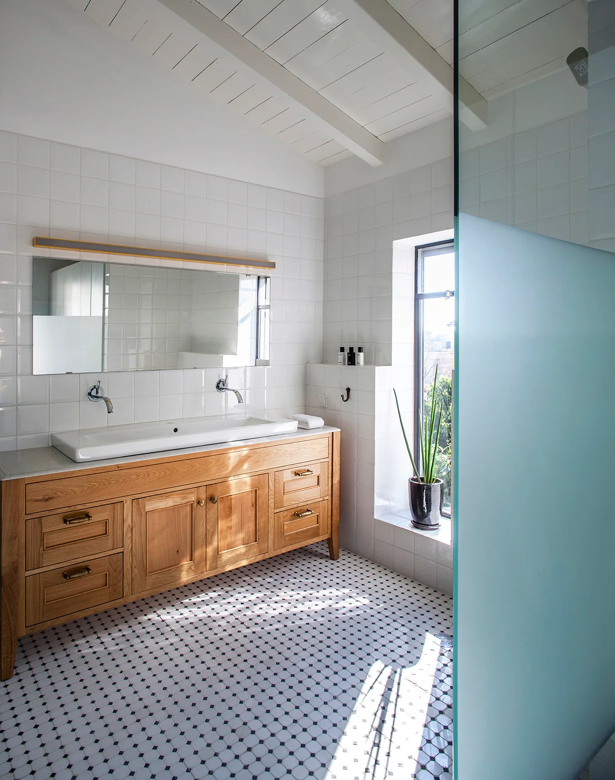 A bright bathroom features a natural wood vanity with double sinks, white tiled walls, and a black-and-white mosaic patterned floor.