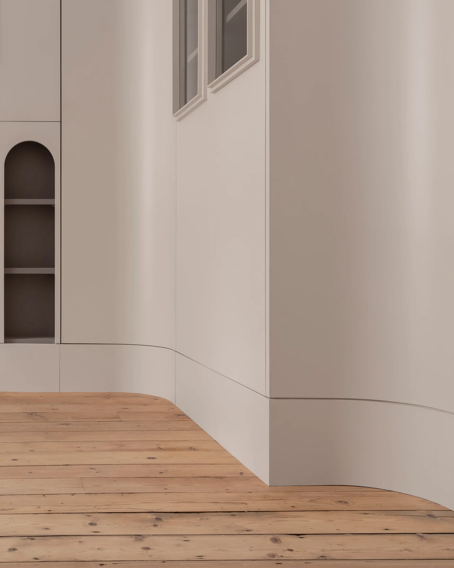 A minimalist room features smooth curved greige walls, a built-in arched shelving unit, and wide plank light wood floors.