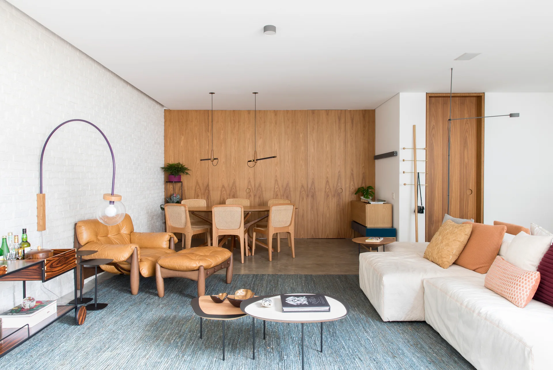 An open-concept living and dining room with a white brick wall, a warm wood paneled accent wall, a cream sectional sofa, and a large blue rug.