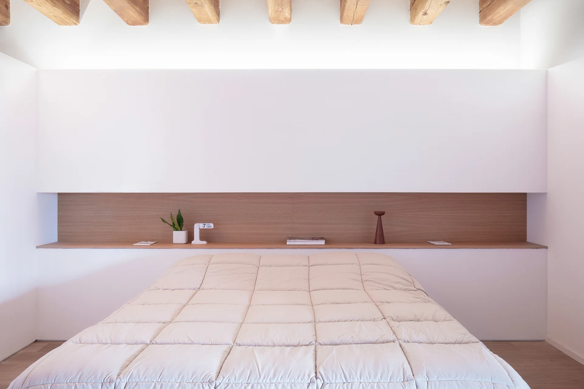 A minimalist bedroom features a bed with a quilted coverlet set against a white wall with a built-in wooden shelf and exposed ceiling beams.