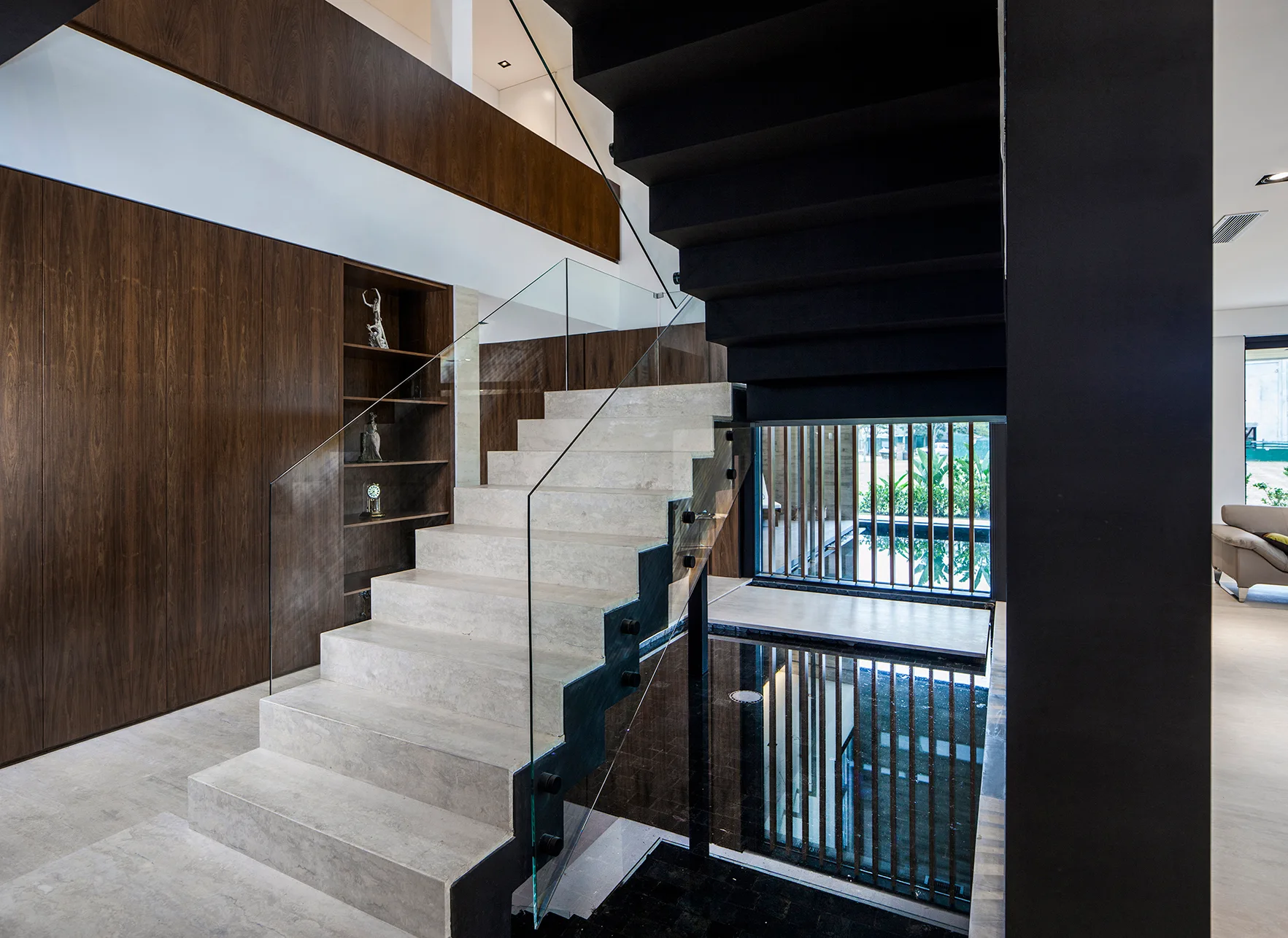 A modern stone staircase with a glass railing ascends over a reflective indoor water basin next to a dark wood paneled wall.
