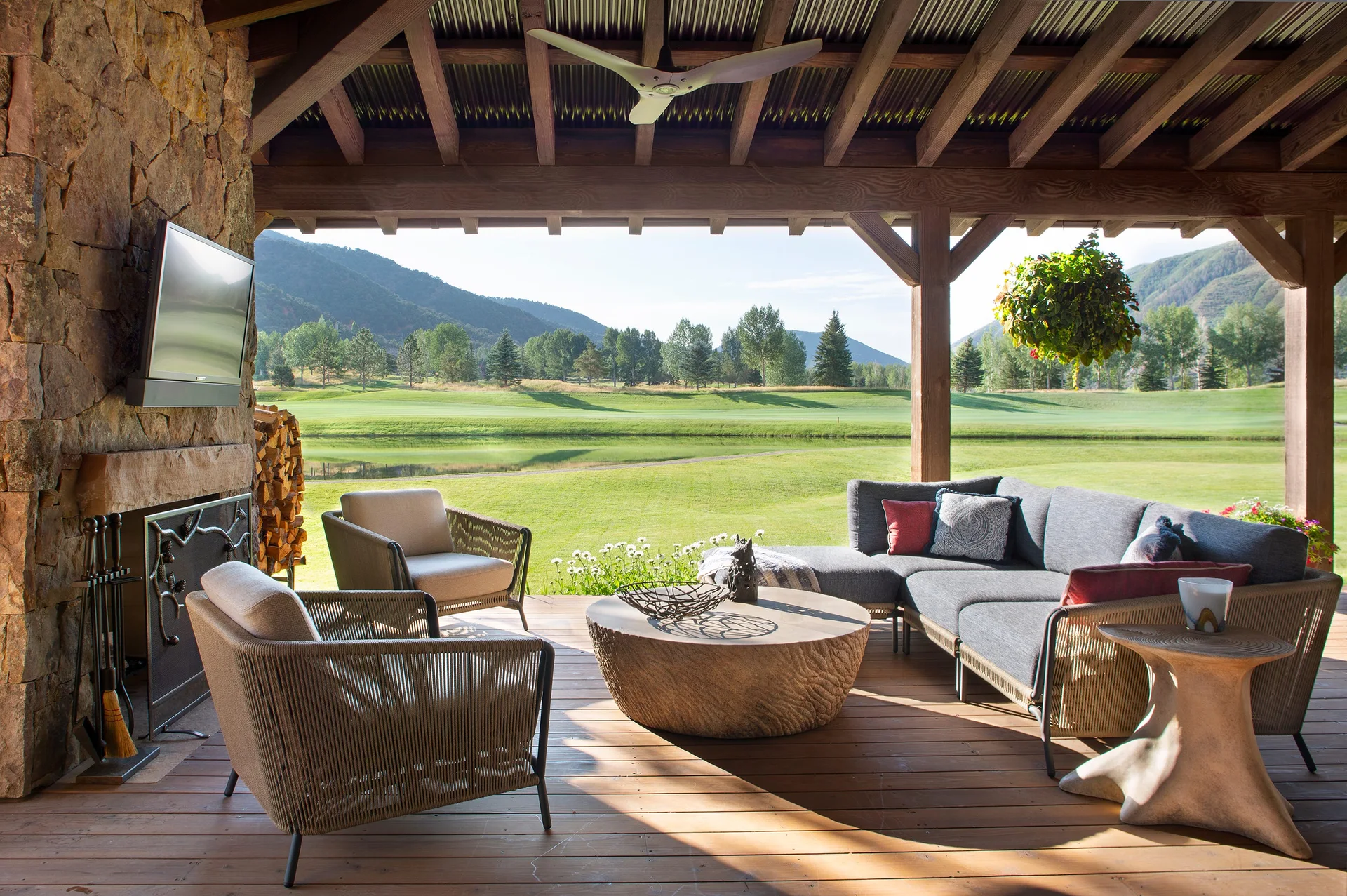 A covered wooden deck features a stone fireplace, a grey sectional sofa, woven armchairs, and an open view of a green landscape with mountains.