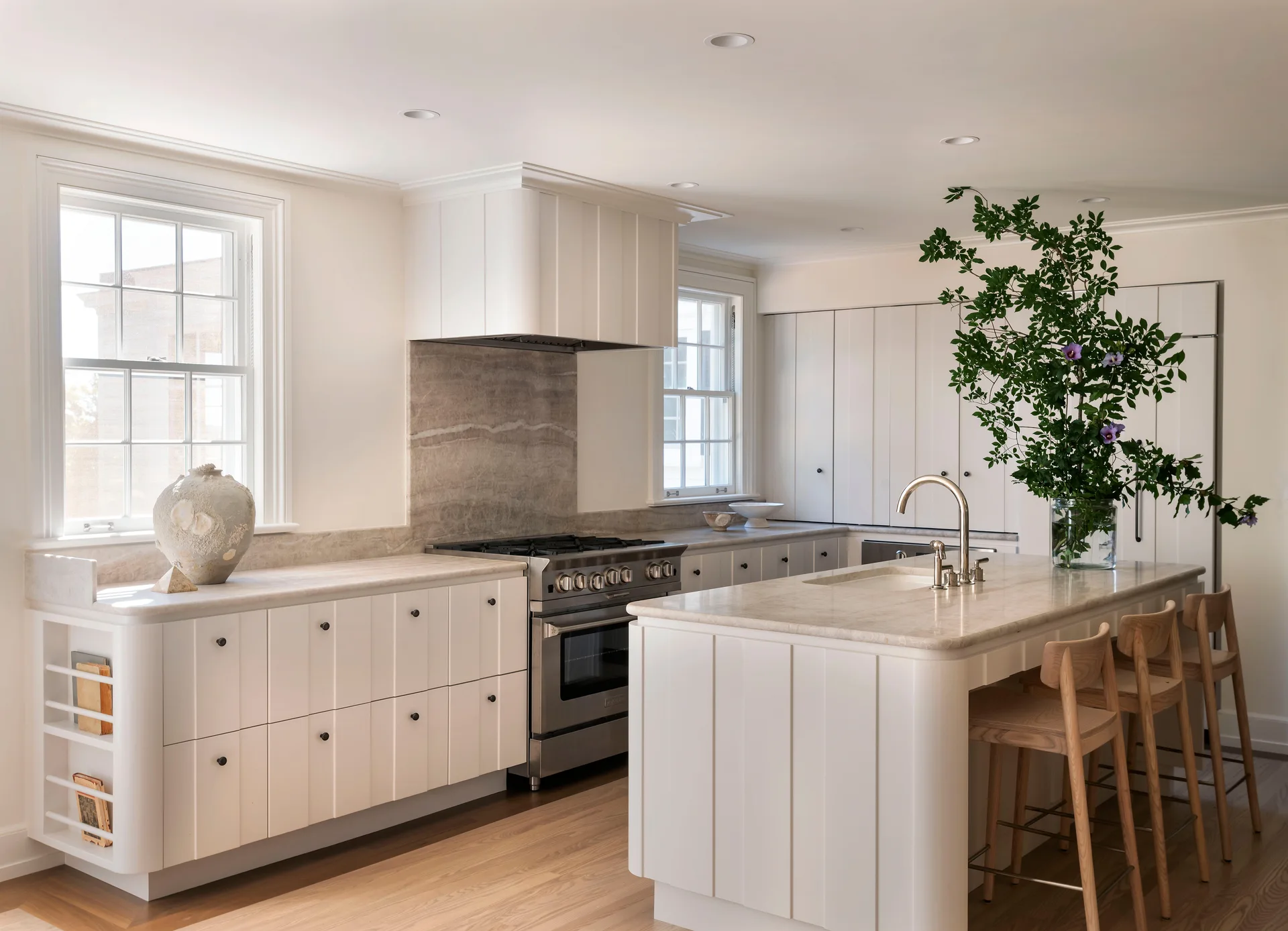 A bright kitchen features off-white vertical panelled cabinets, a central island with wood stools, and a stone countertop and backsplash.