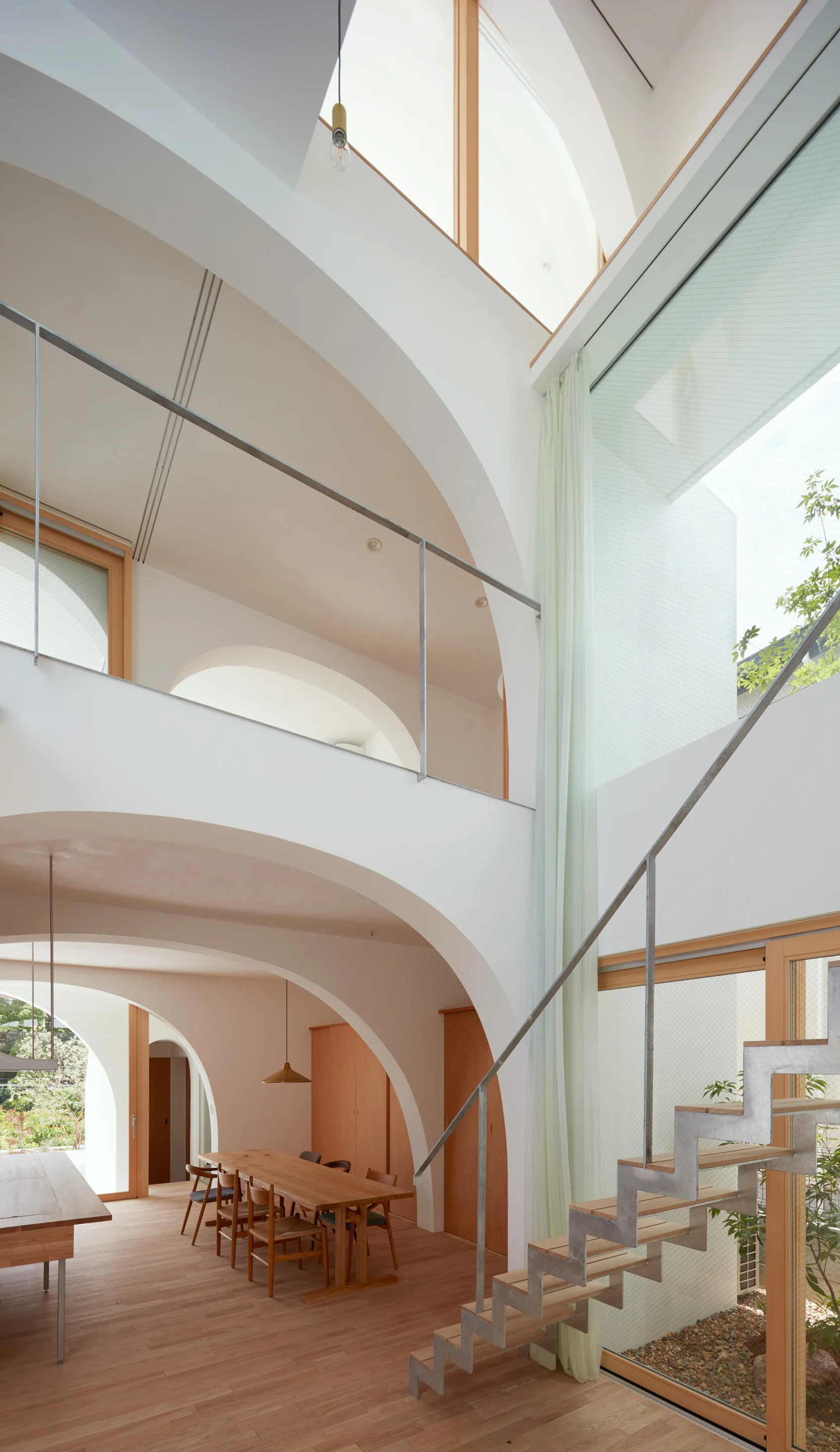 A bright, multi-level dining space features large white architectural arches, a long wooden dining table, and an open metal staircase with wood steps.