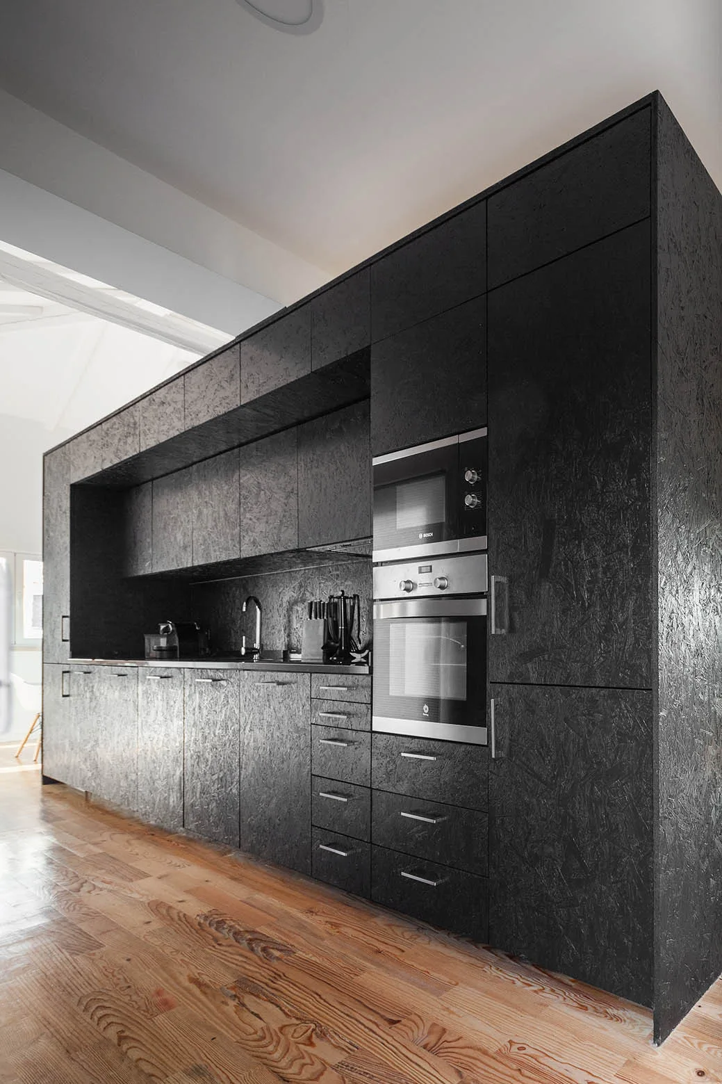 A modern kitchen featuring a full wall of textured black OSB cabinetry with built-in steel appliances on a warm wood floor.