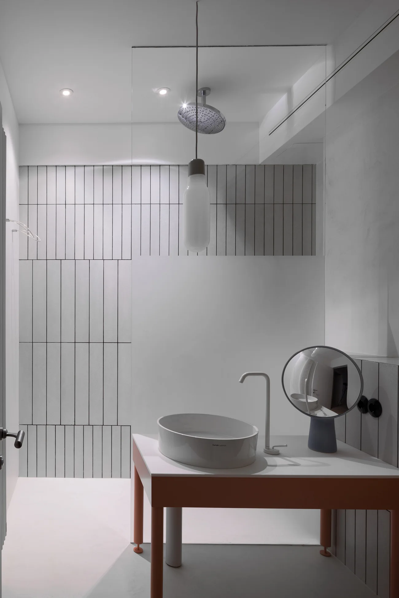 A minimalist bathroom features a coral-framed vanity with an oval vessel sink, vertical gray-and-white wall tiles, and a hanging pendant light.
