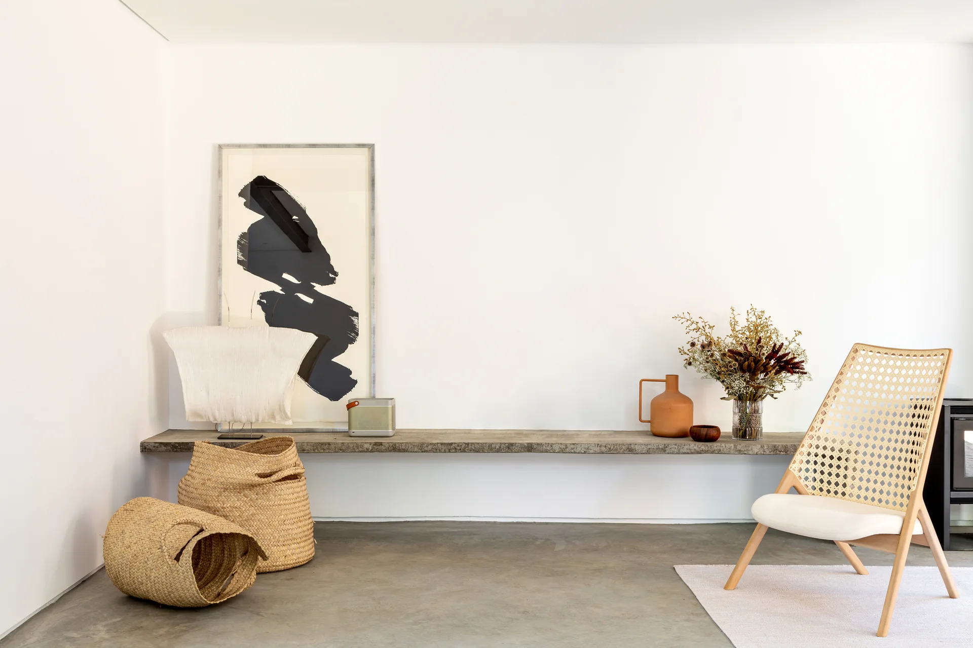 A minimalist room featuring a long floating concrete shelf adorned with artwork and vases, a cane accent chair, and woven floor baskets.