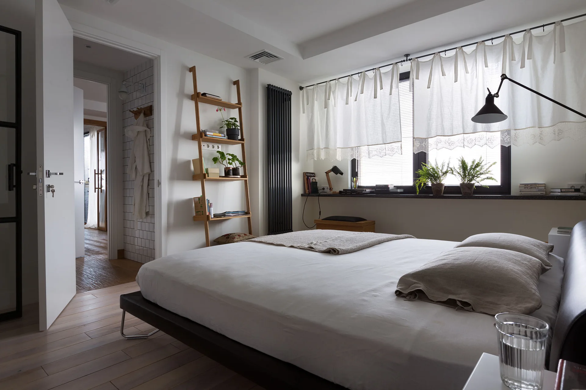 A modern bedroom featuring a low-profile bed, a leaning wooden ladder shelf, a black vertical radiator, and sheer curtains over the window.