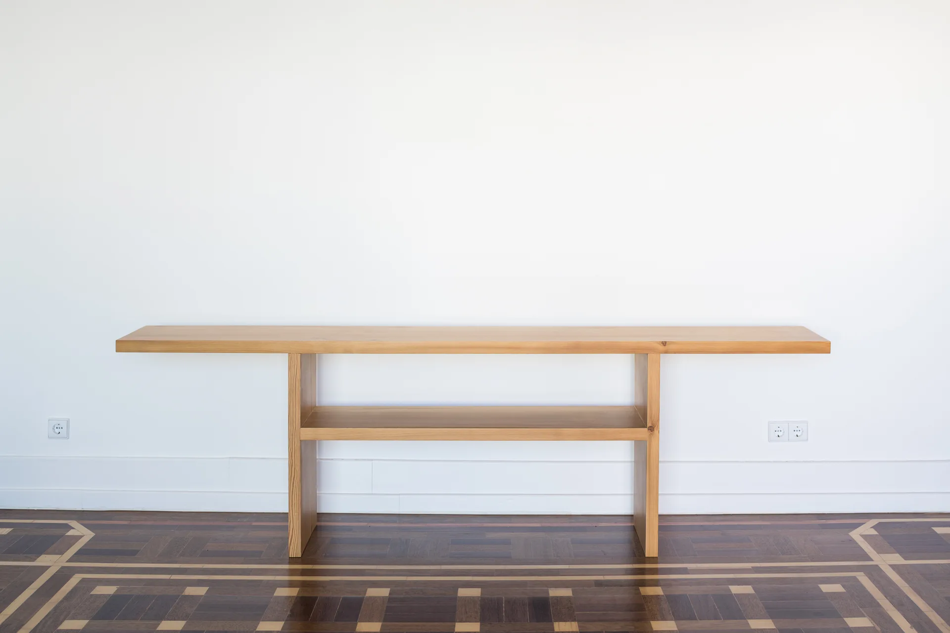 A minimalist wooden console table with an extended top stands against a white wall on a dark geometric parquet floor.