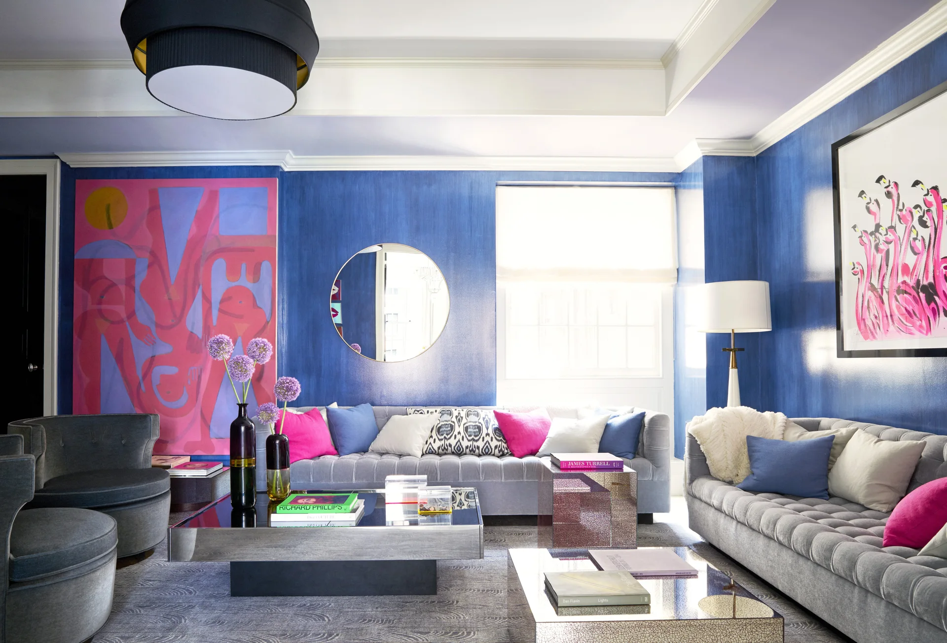 A living room features brushed blue walls, two gray tufted sofas, colorful pink and purple throw pillows, and reflective coffee tables.