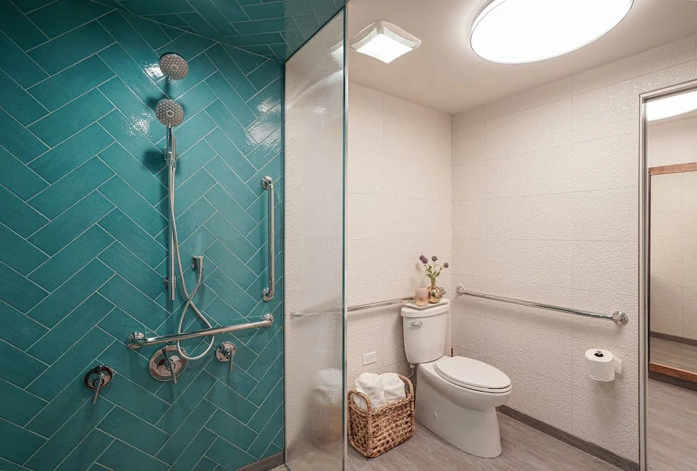 A modern bathroom featuring a glass-enclosed shower with teal herringbone tile next to a white toilet with stainless steel grab bars.