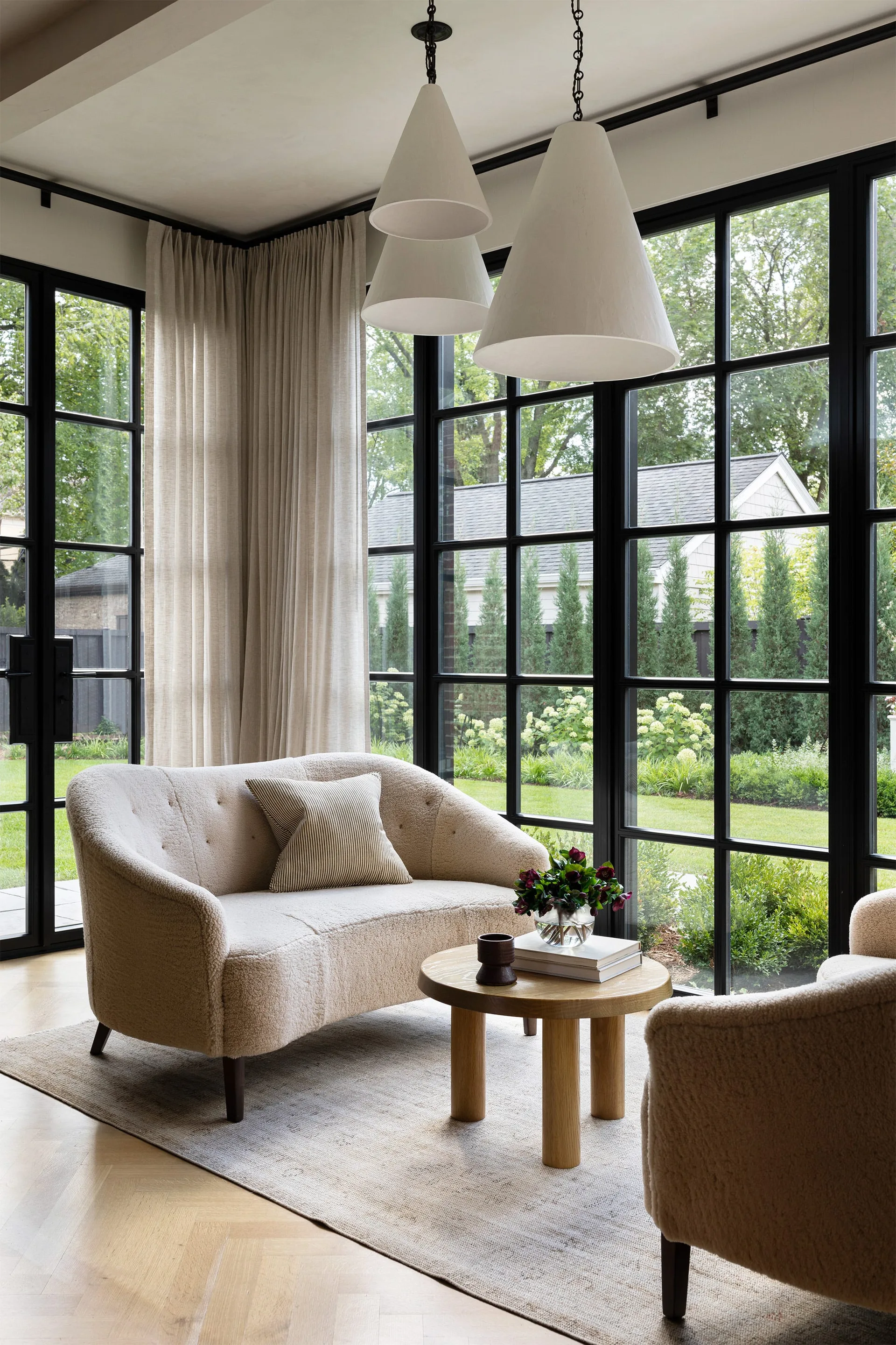 A bright living space features a curved beige bouclé sofa, a small round wooden coffee table, and large black grid windows looking out to a garden.
