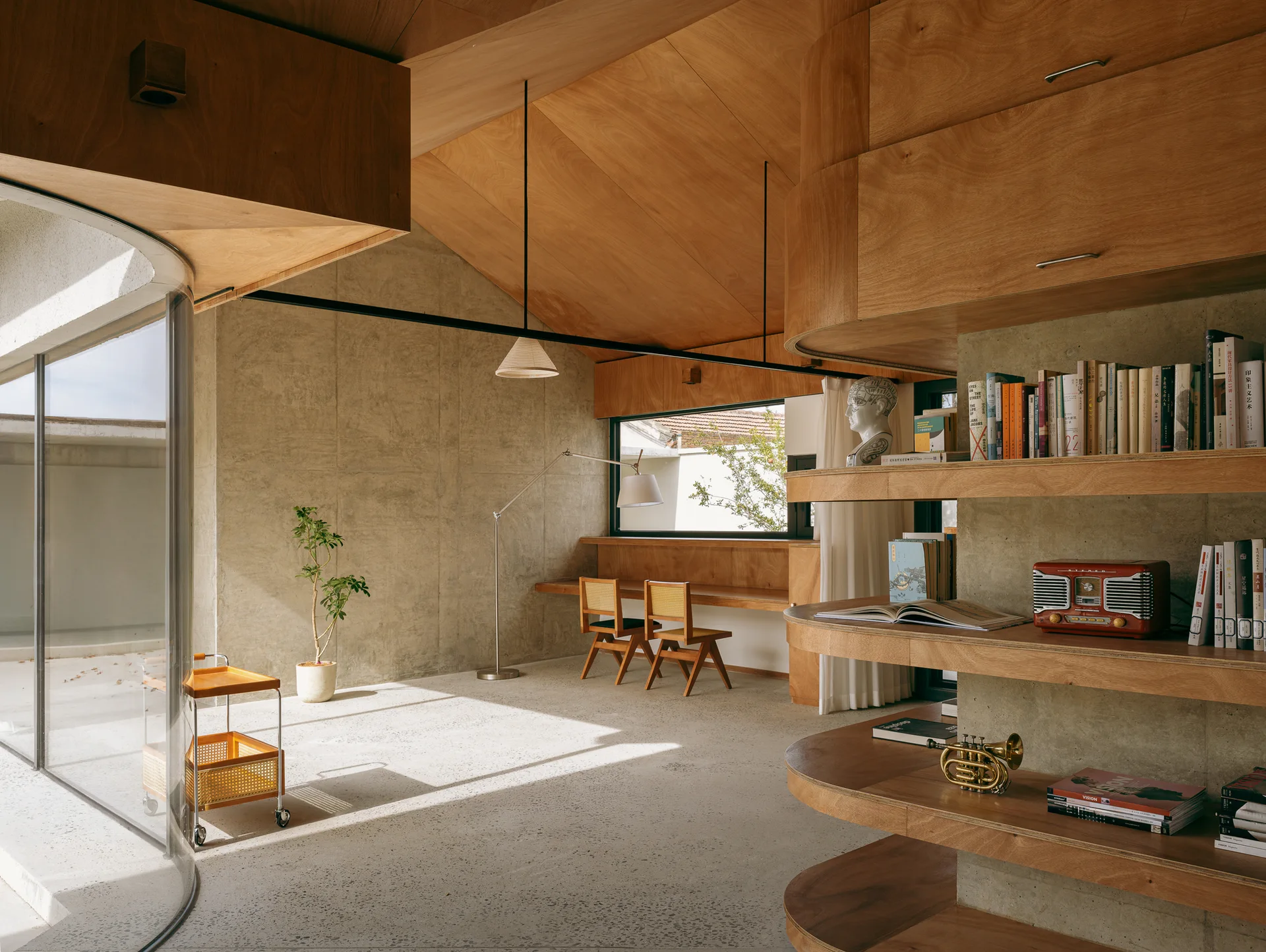 A modern room features a sloped plywood ceiling, a curved wood shelving unit, raw concrete walls, a terrazzo floor, and a built-in desk.