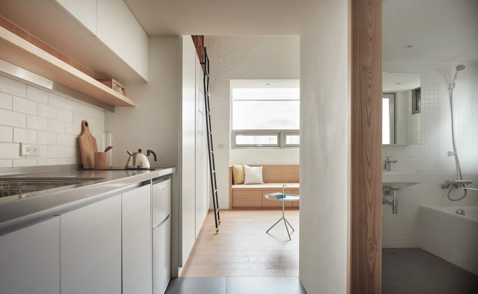 A compact modern apartment view looking from a white-cabinet kitchenette toward a wooden window bench with a black rolling ladder.