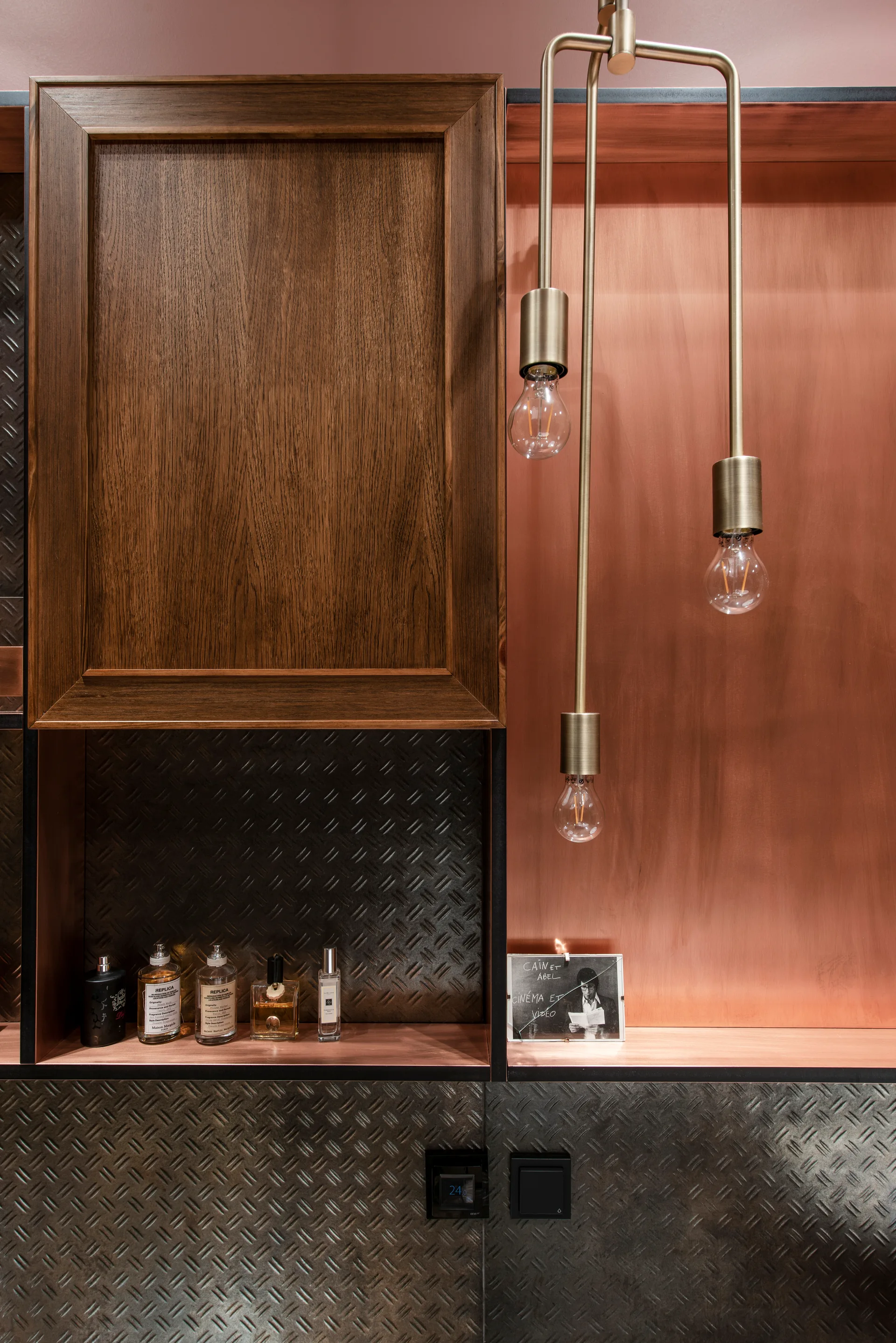 A close-up of a wood cabinet, a copper-backed open shelf with perfume bottles, and a brass pendant light against dark diamond-plate wall paneling.