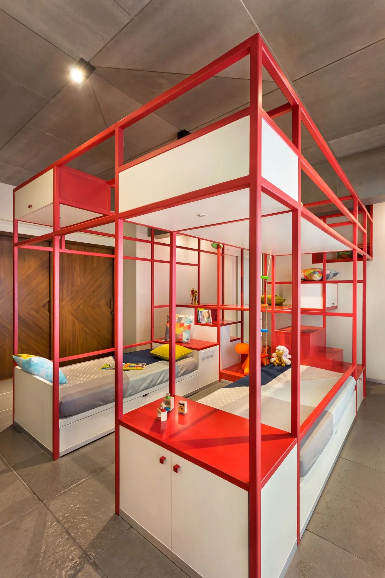 A modern children's bedroom features a custom bunk bed unit with a bright red metal frame, integrated storage cabinets, and concrete ceiling.
