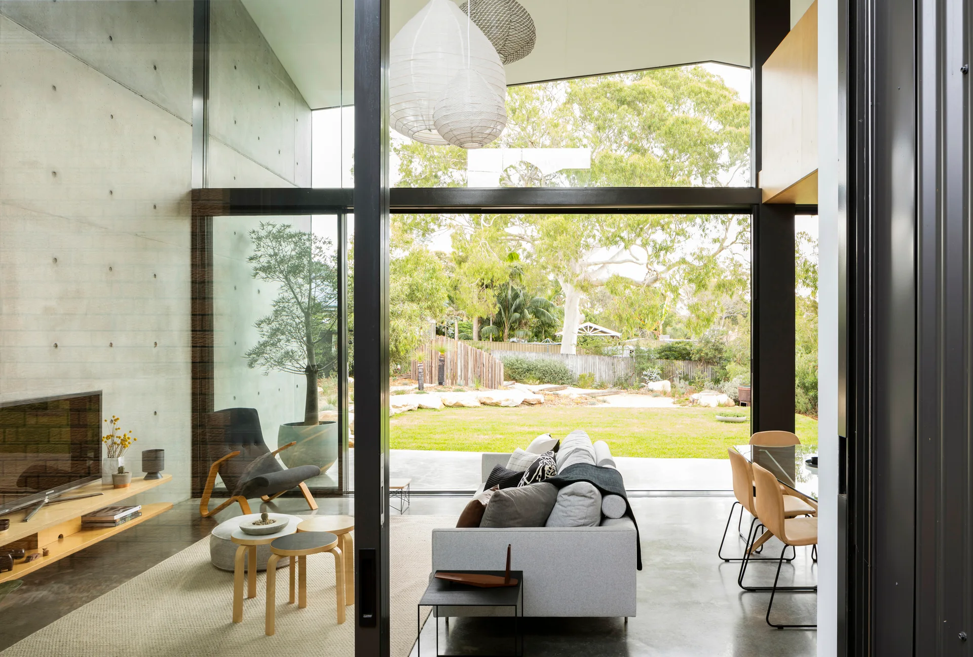 A modern living room features an exposed concrete wall, a gray sofa, nested wood tables, and a large sliding glass door opening to a grassy backyard.