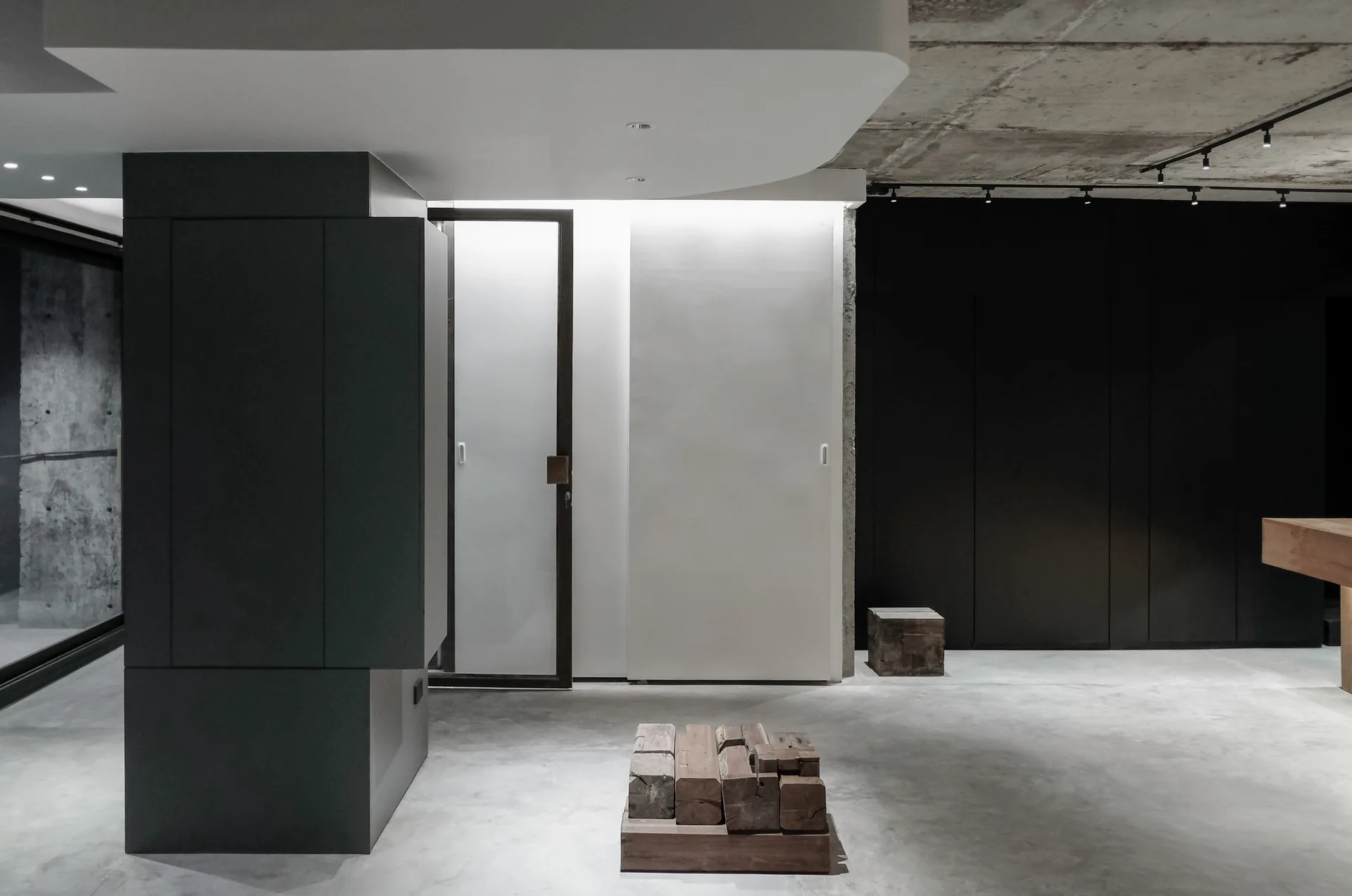 A minimalist interior featuring a raw concrete ceiling, dark gray cabinetry, a white dropped ceiling, and a wooden block sculpture on a concrete floor.