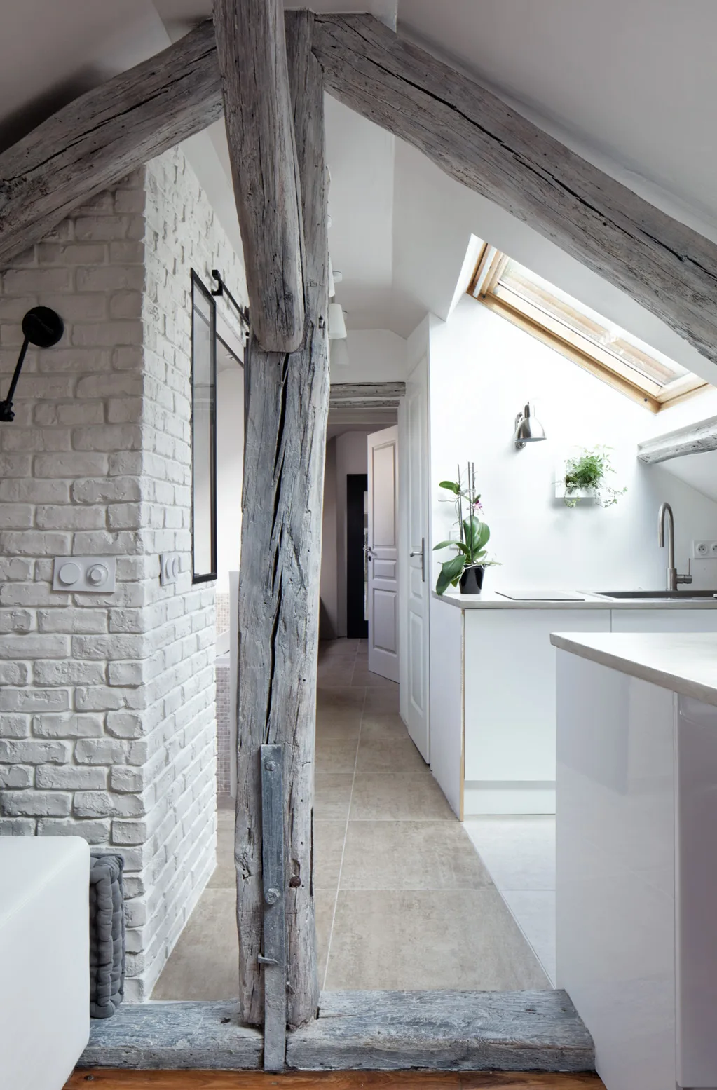 A bright attic kitchen features clean white cabinetry, a skylight, a white painted brick wall, and prominent rustic wood timber beams.