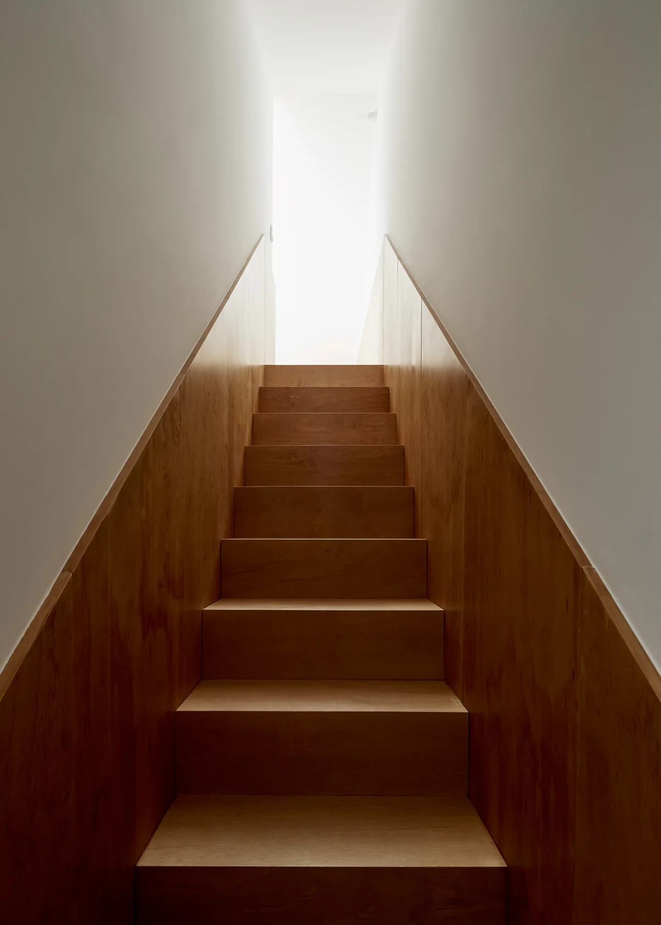 A straight wooden staircase with warm wood-grained paneling on the walls leads up toward a bright light.