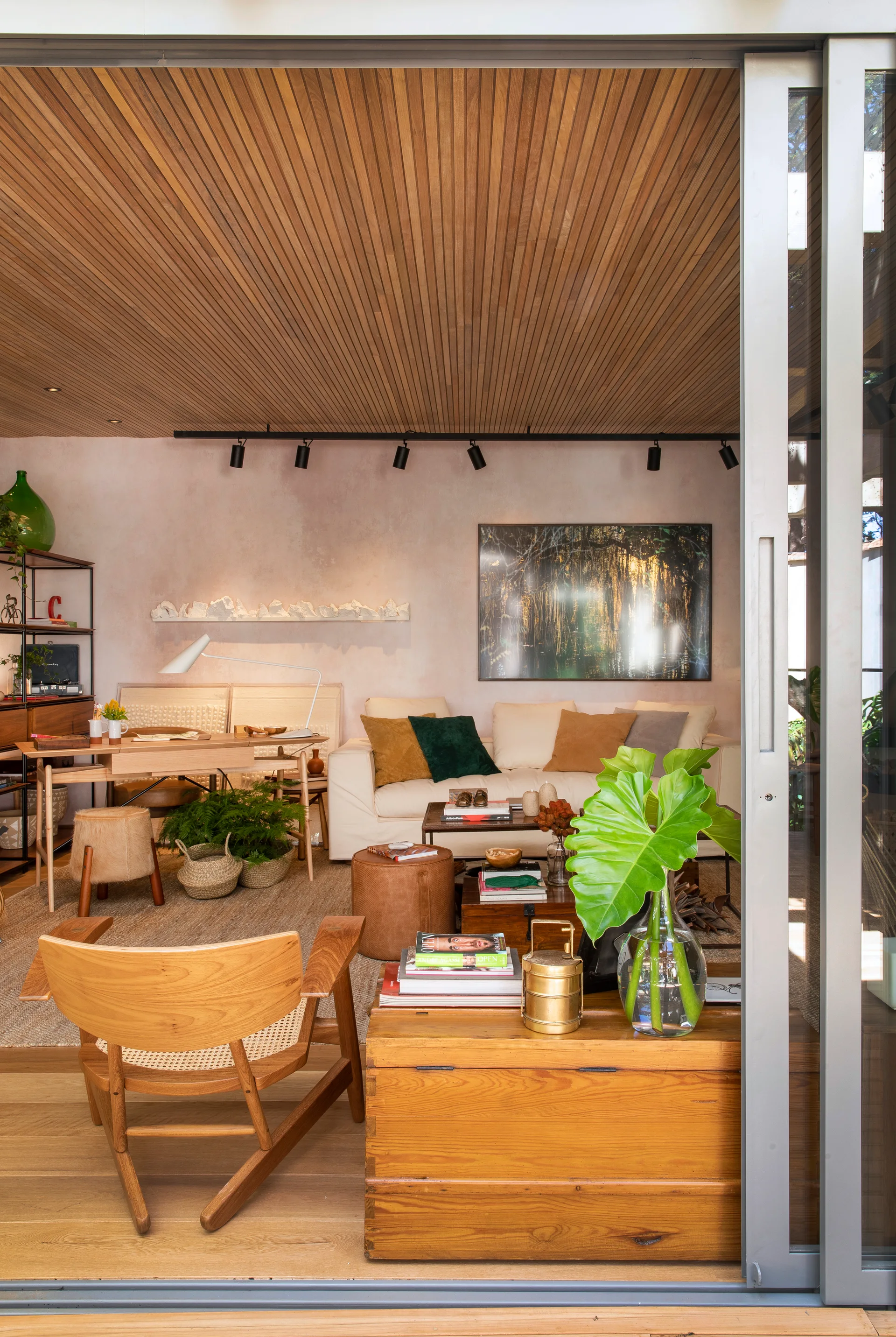 An organic modern living room featuring a slatted wood ceiling, a cream sofa, and a wooden desk, viewed through sliding glass doors.