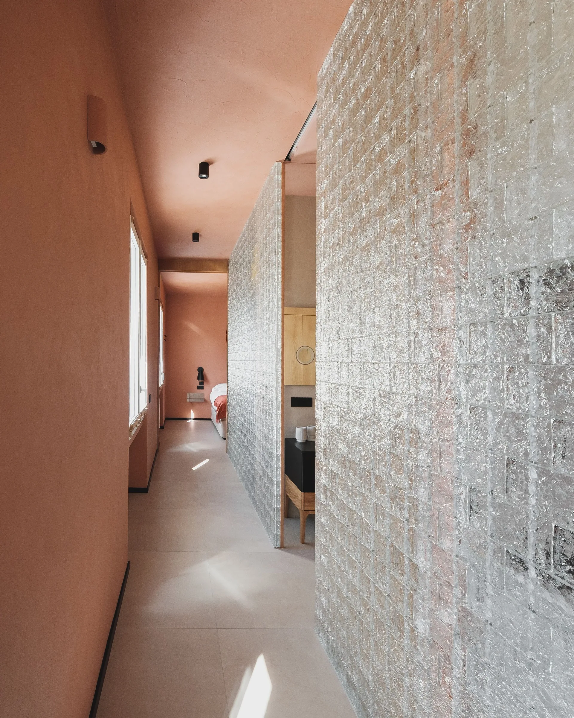 A long hallway with matte pink plaster walls and ceiling alongside a translucent textured glass block partition wall.