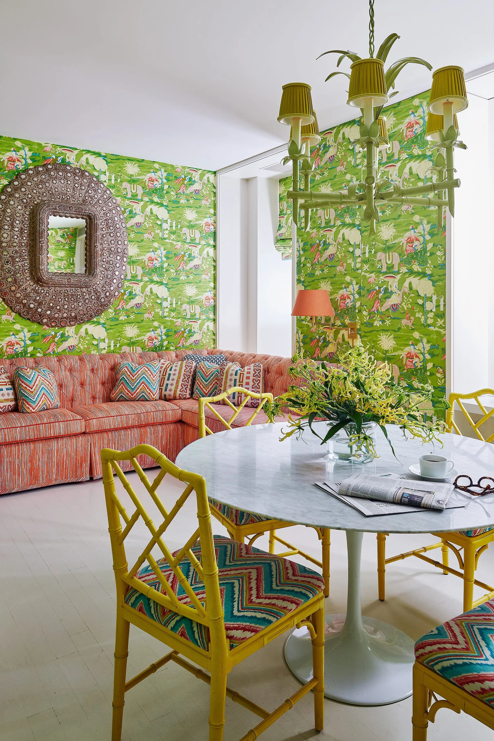 A dining area features yellow bamboo chairs and a marble table set against green patterned wallpaper and an orange tufted sectional sofa.