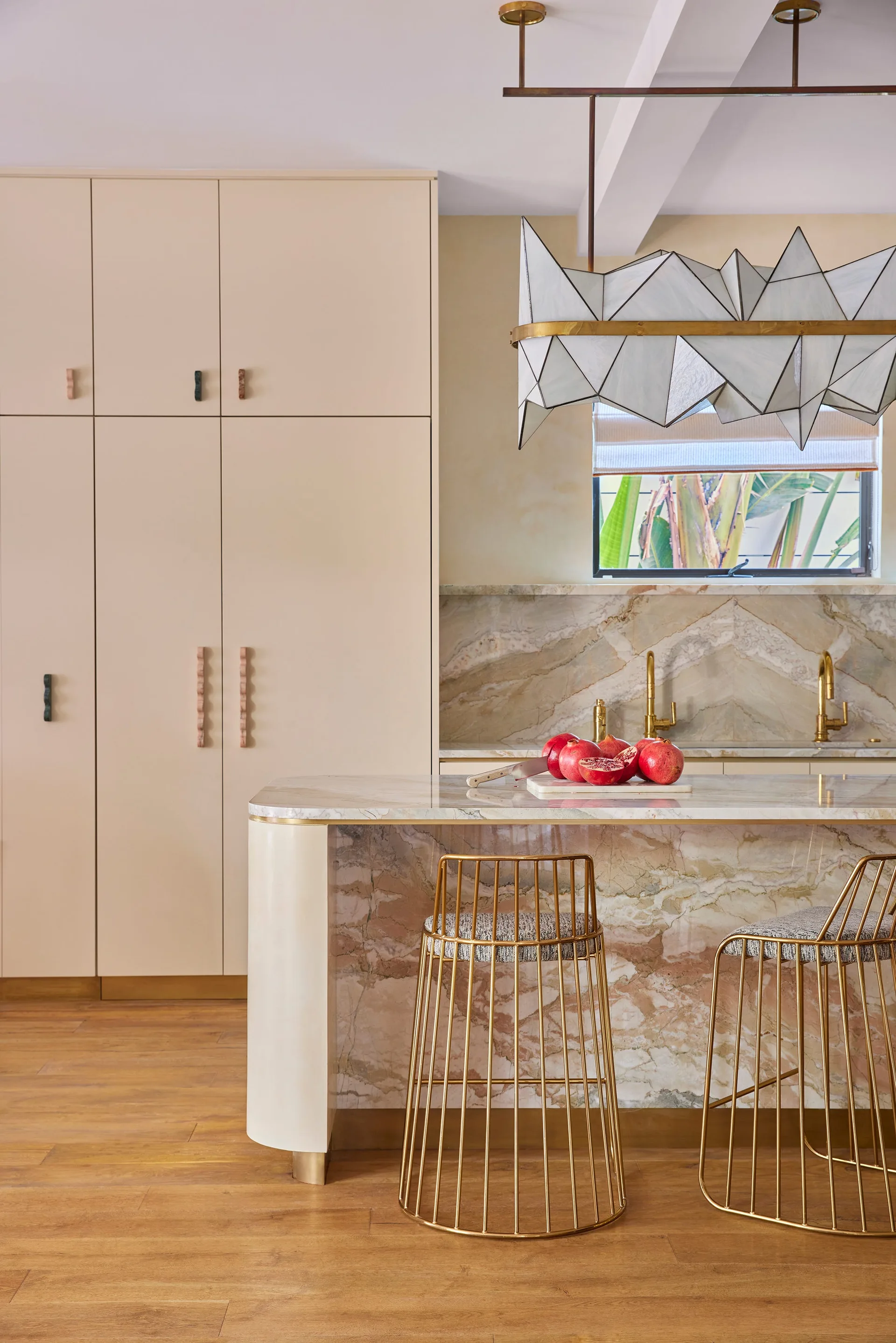 A modern kitchen showing a marble-clad island with two brass wire-frame bar stools, pale cream cabinetry, and a geometric pendant light.