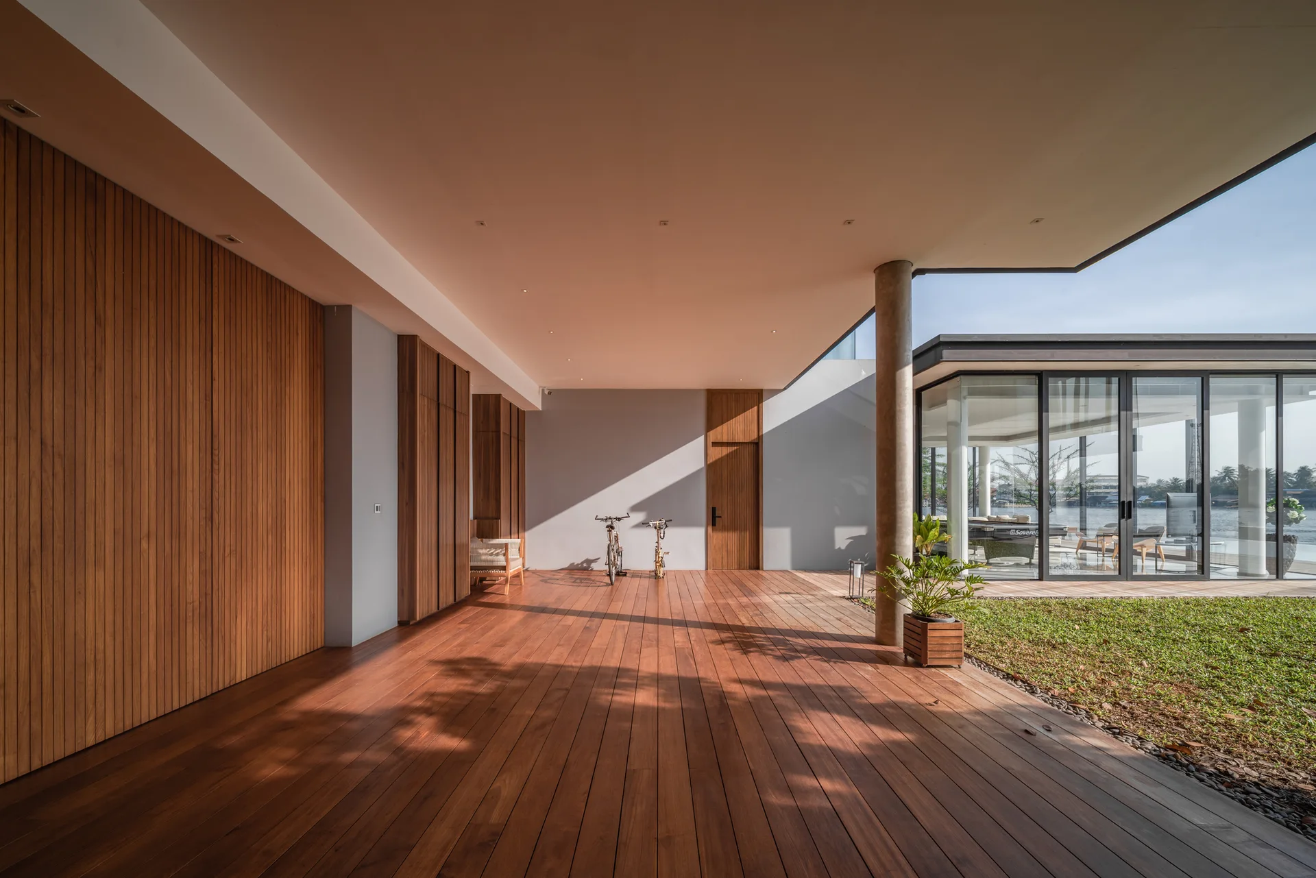 A spacious covered patio features warm-toned wooden decking, vertical slatted wood wall cladding, a concrete column, and a grassy garden nearby.