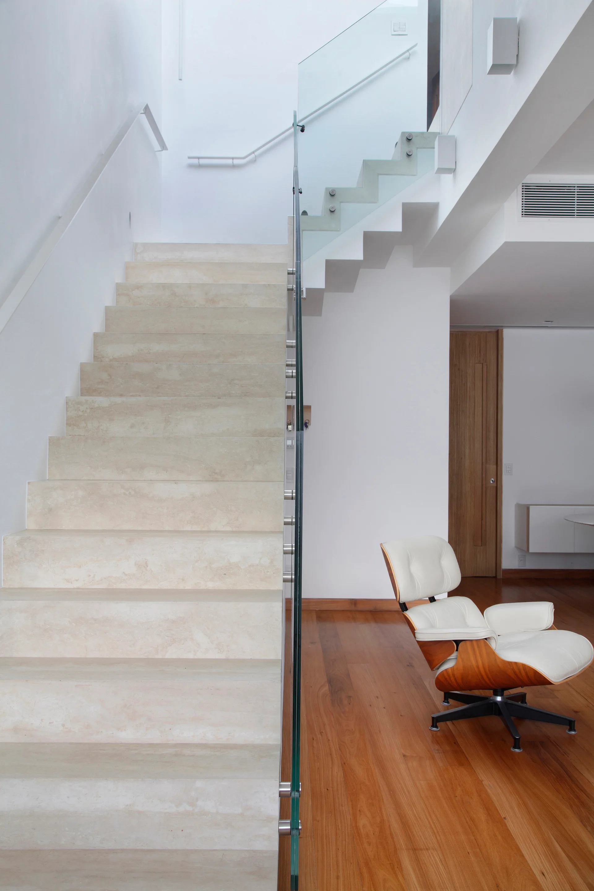 A modern stone staircase with a clear glass railing ascends next to a white leather lounge chair on warm hardwood floors.
